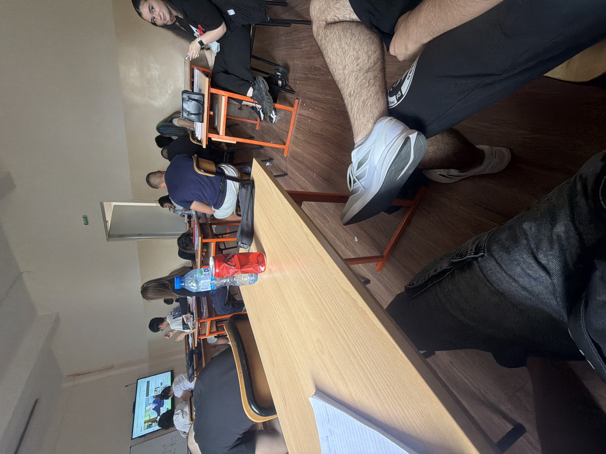 The image depicts a classroom scene with multiple students seated at desks. A large screen is mounted on the wall, displaying content that appears to be a video lecture or presentation. Several students are facing the screen, some paying attention, while others are looking away or engaged in other activities. In the foreground, the legs and feet of two individuals are visible, one wearing jeans and the other black pants with a white logo. A wooden desk occupies a significant portion of the foreground, with a notebook and a crushed red can of Coca-Cola on it. There are water bottles and other personal belongings scattered on desks throughout the classroom. The classroom environment includes fluorescent lighting and simple wooden desks and chairs. The presence of an "EXIT" sign above a doorway suggests this is a public building. Without further context, the specific location within Sofia, Bulgaria cannot be determined.