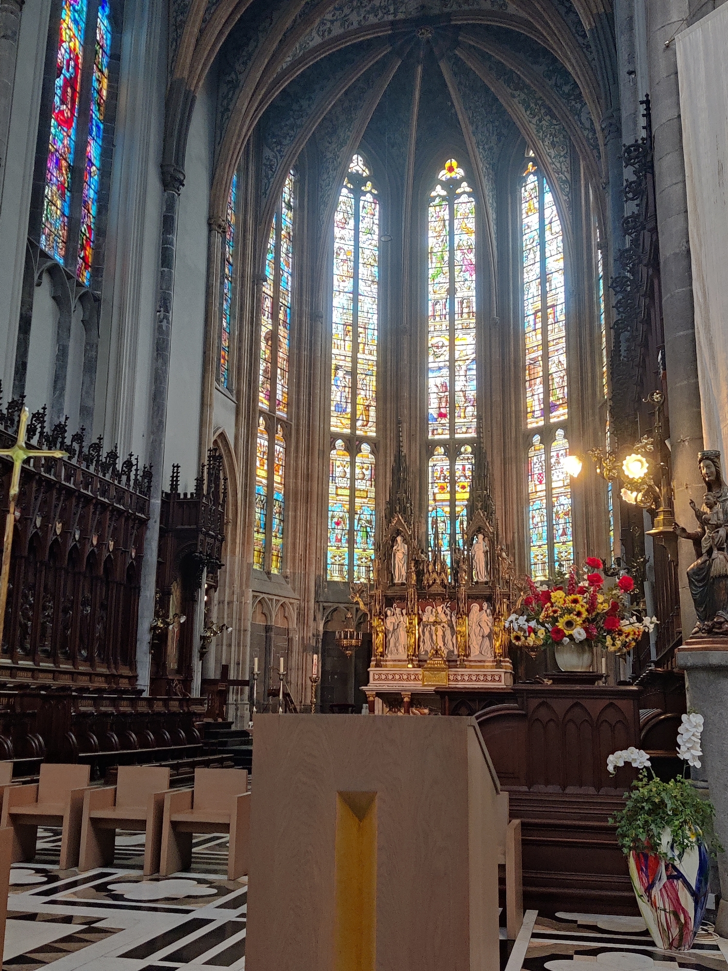 The image shows the interior of a Catholic church in Liège, Belgium, specifically Saint-Jacques-le-Majeur. Sunlight streams through a large, ornate stained-glass window at the far end of the nave, illuminating the space with vibrant colors. The window features numerous panels depicting religious scenes and figures.
In the foreground, a wooden pulpit with a gold-colored accent stands at an angle. To the left of the pulpit are several modern, light-brown chairs arranged in rows on a patterned floor with geometric designs. A wooden confessional and choir stalls, carved with intricate details, line the sides of the nave. A golden cross is affixed to the side of the choir stalls.
The altar area is adorned with several statues of religious figures and a large floral arrangement of sunflowers, red and yellow roses, and white flowers in a vase. To the right of the altar, a dark wooden sculpture of a woman holding a child stands on a pedestal. A potted plant with white orchids and green foliage is placed near the front of the altar. The overall environment is one of grandeur and solemnity, with a blend of ancient architecture and contemporary furnishings.