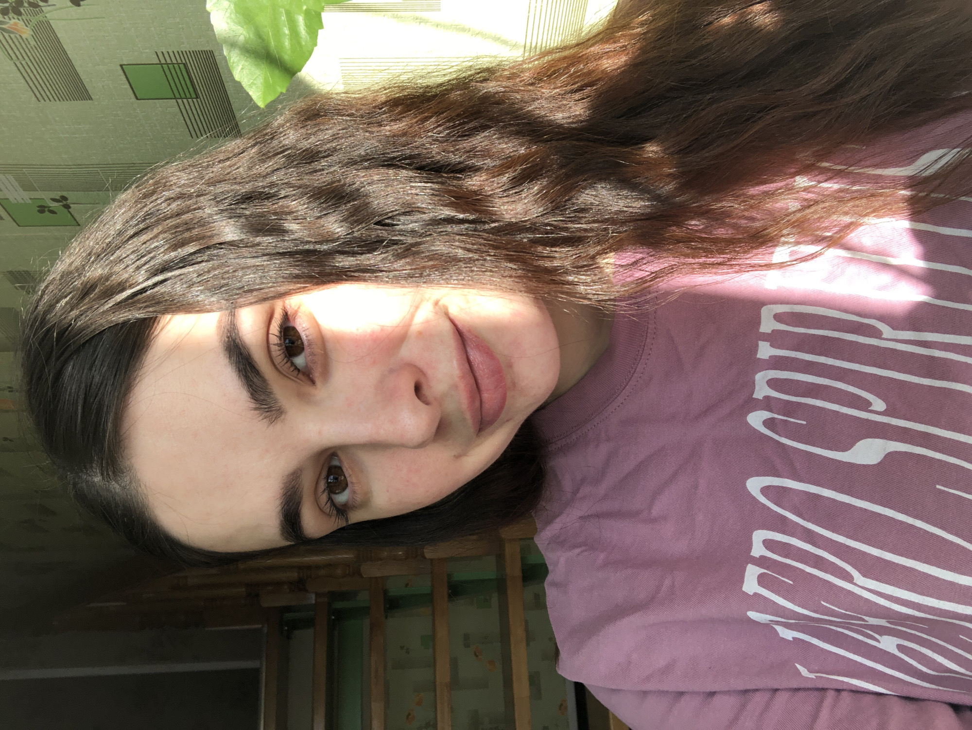 A young woman with long, dark brown wavy hair is posing indoors. She is fair-skinned with dark eyes and thick eyebrows. She's wearing a mauve or light purple t-shirt with white lettering partially visible. The background includes a green and brown patterned surface (possibly wallpaper) and a glimpse of bright sunlight. The photo appears to have been taken at an angle. There is no indication of Noviy, Russia in the picture.