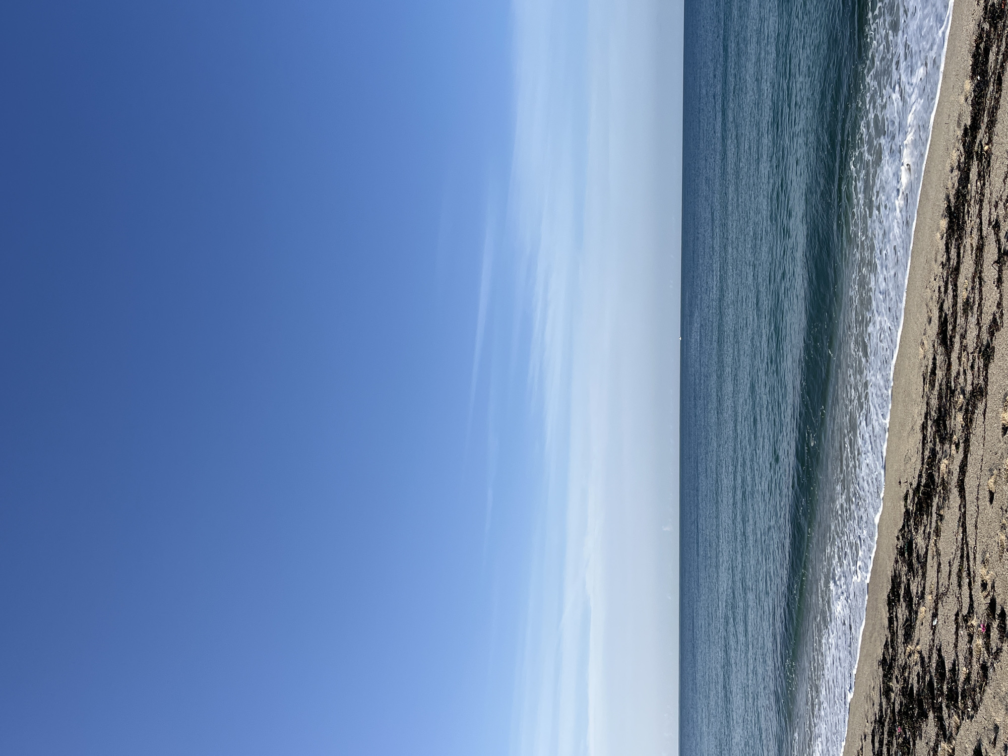 The image depicts a beach scene with the sky occupying the majority of the left side. A clear blue sky fades into a hazy, lighter blue towards the center. The right side shows the ocean and the beach. The water is a deep blue color, with white foamy waves reaching the shore. The beach itself is sandy and covered in dark seaweed or debris. The scene appears to be taken on a bright, sunny day. The location is possibly a beach in Alanya, Turkey, as requested.
