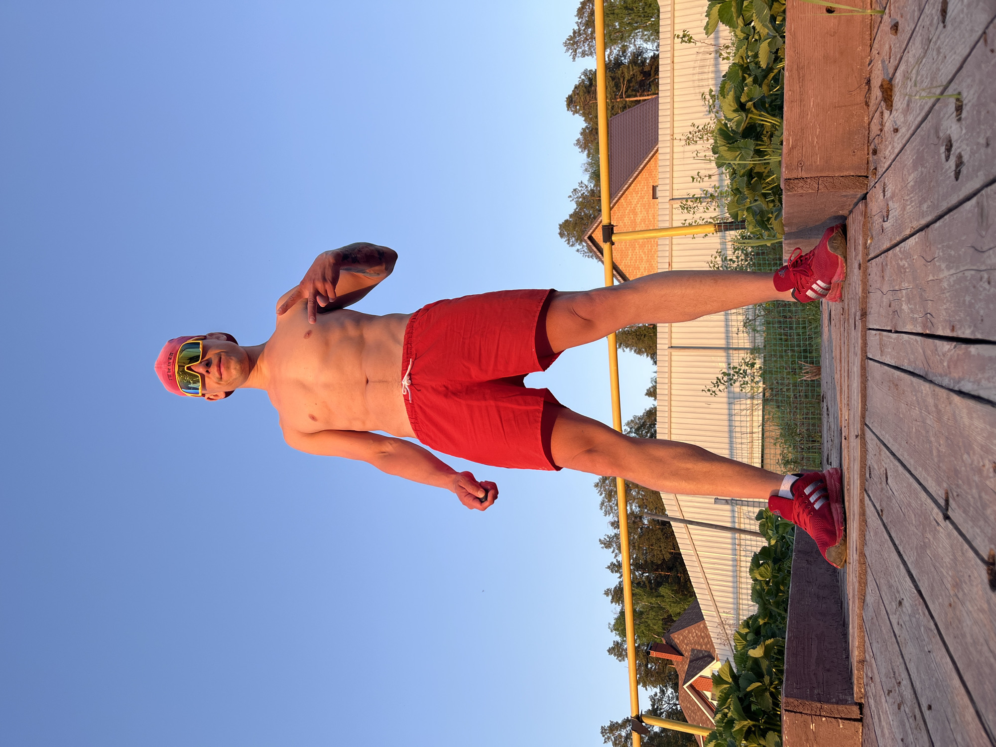 A shirtless man wearing red shorts, red shoes with white stripes, a pink cap, and yellow sunglasses stands on a wooden surface outdoors. He has a tattoo on his upper arm. The background includes a blue sky, a yellow fence, a building with a brown roof, and greenery. The location is identified as the city of Chaikovsky, Russia.