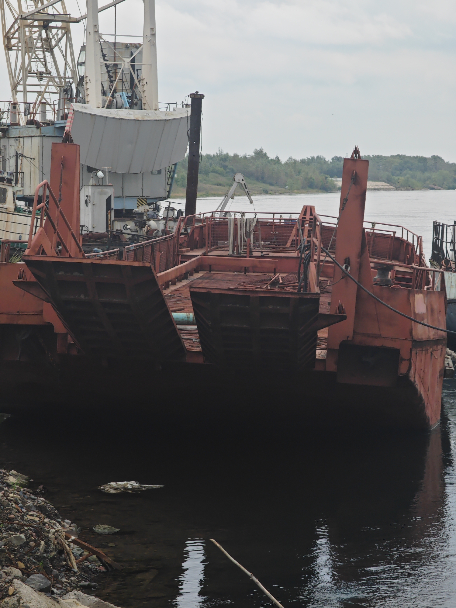 An old, rusty red barge is docked on the bank of a wide river, possibly the Yenisei River in Krasnoyarsk, Russia. The barge is large and appears to be a ferry or cargo vessel, with a wide, flat deck and ramp-like structures at the front. Behind the barge, another vessel, possibly a crane ship, is partially visible, suggesting it might be part of a shipyard or salvage operation. The water is dark and reflects the overcast sky. The riverbank on the right side of the frame is lined with trees, indicating a natural landscape beyond the industrial setting. The overall impression is one of disuse and decay, with the barge seemingly abandoned or awaiting repairs.
