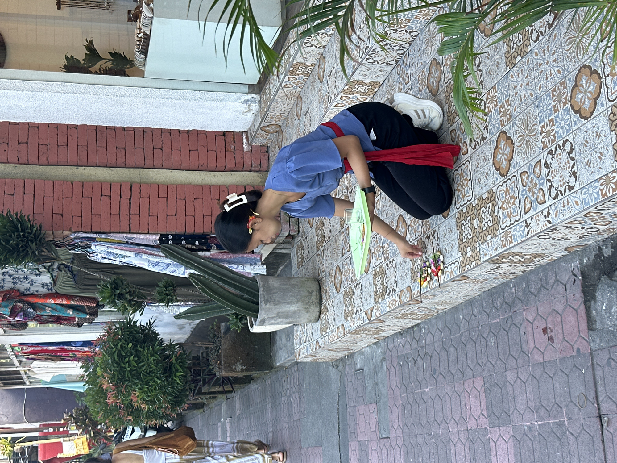 A young woman with dark hair, wearing a blue shirt, black pants, and a red sash, kneels on a tiled step outside a shop in Ubud, Indonesia. She is placing small, colorful offerings on the step. In front of her is a green tray with a glass of water. The shop displays clothing and plants. A person in a white top and patterned pants walks by in the background. The street is paved with interlocking bricks.
