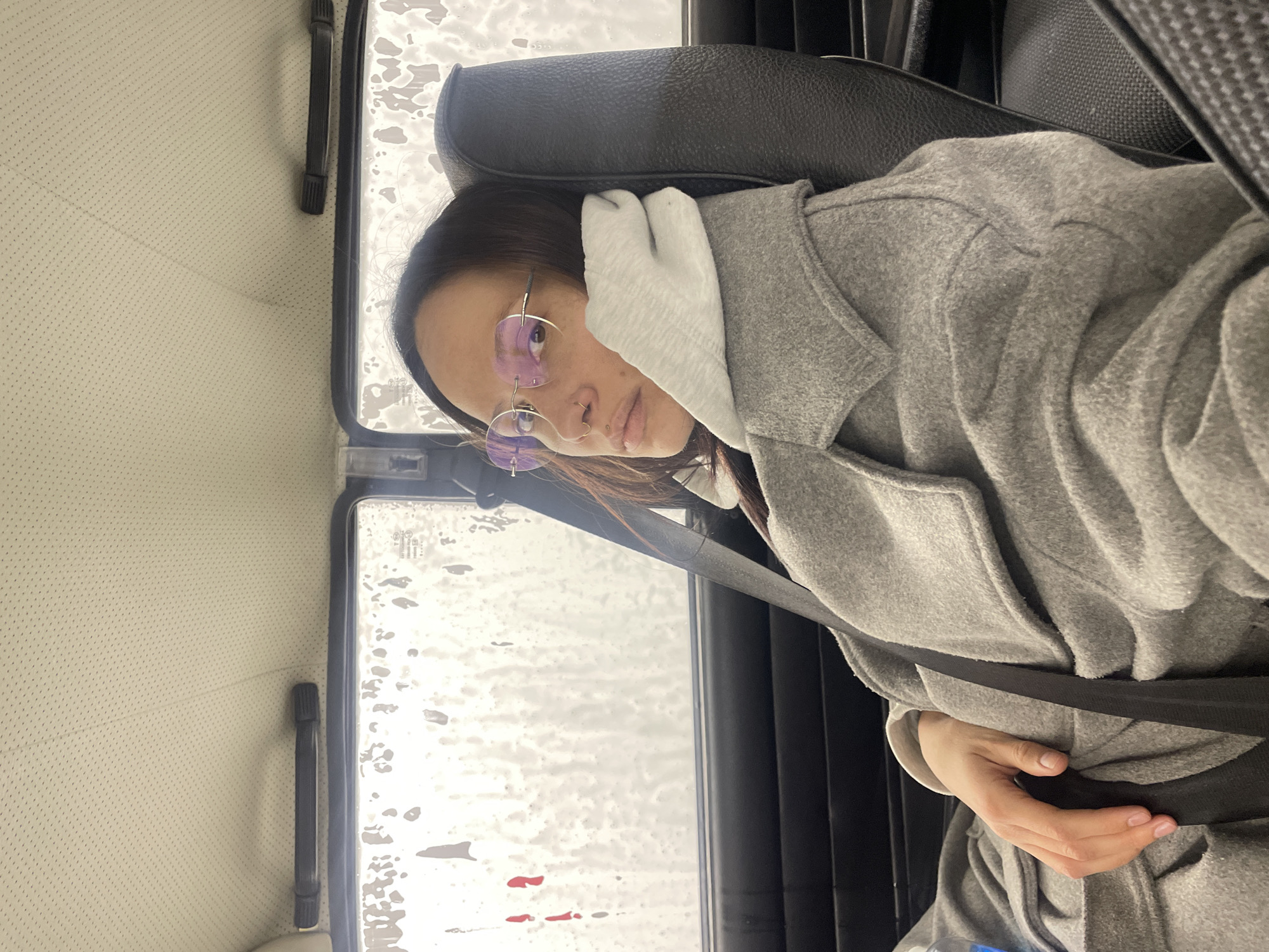 A person is seated in a vehicle, likely a car, wearing a gray coat and a light-colored hooded sweatshirt. They are wearing glasses with light-colored frames. The seatbelt is fastened. The window is covered in soapy water or foam, possibly in a car wash. The interior of the car appears to be somewhat vintage, with a light-colored, perforated headliner. There is no verifiable evidence to indicate that the location is in the city of Izhevsk, Russia.