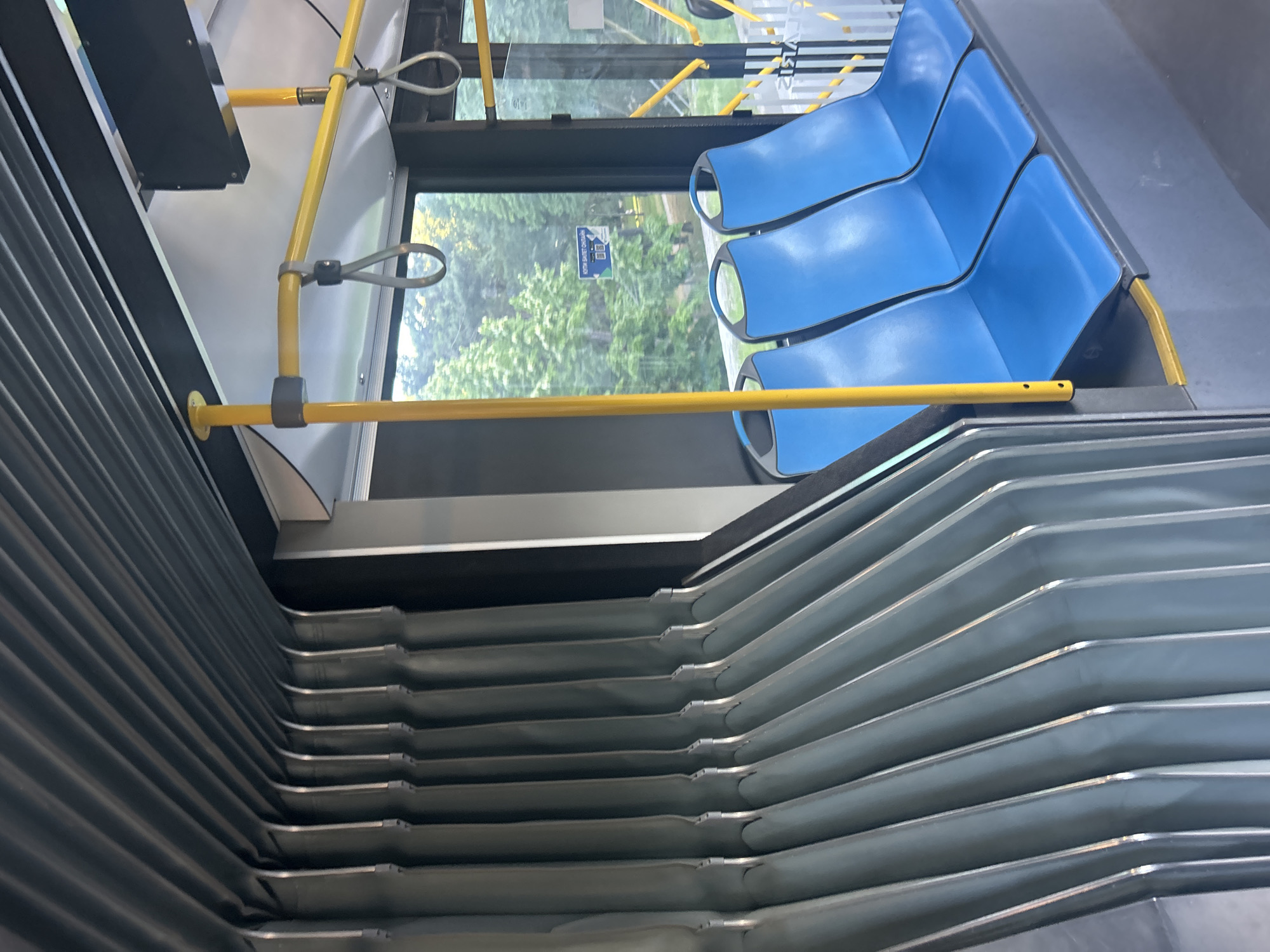 The image shows the interior of a bus, likely a public transport vehicle, with a focus on the seating area and the flexible bellows of an articulated bus. The seats are bright blue and arranged in rows. Yellow poles and grab handles are visible, along with the ribbed, dark gray material of the accordion-style joint characteristic of articulated buses. Through the windows, trees and foliage can be seen, suggesting the bus is in motion or parked near a green area. There are no people visible in the frame.