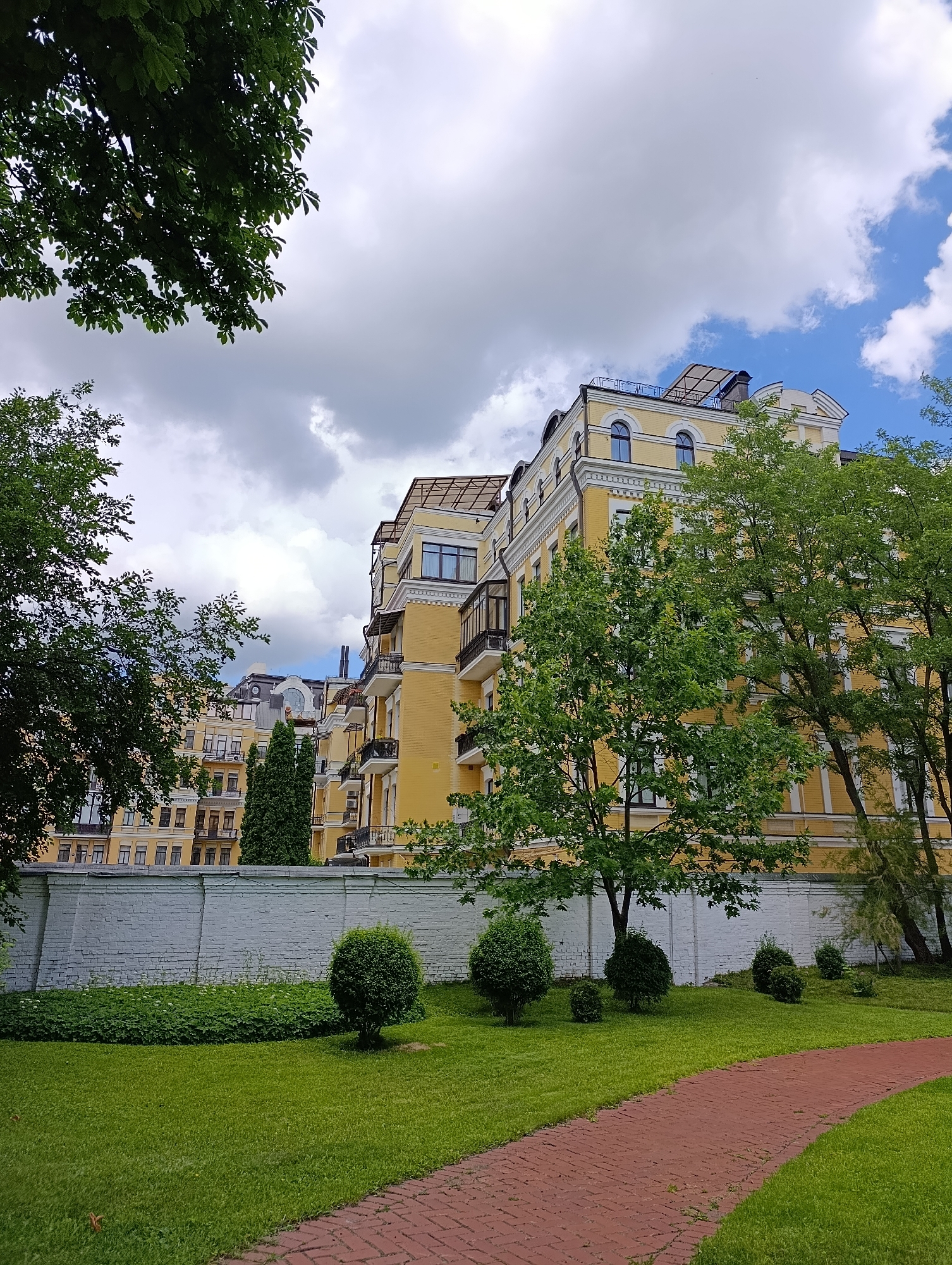 This image shows a view of a modern yellow apartment building in Kyiv, Ukraine, on a cloudy day. The building has multiple floors with balconies and arched windows. In front of the building, there is a white brick wall, a neatly trimmed lawn, several round bushes, and a winding brick path. Trees with green foliage are visible in the foreground and around the building. The sky is partly cloudy with patches of blue sky visible.