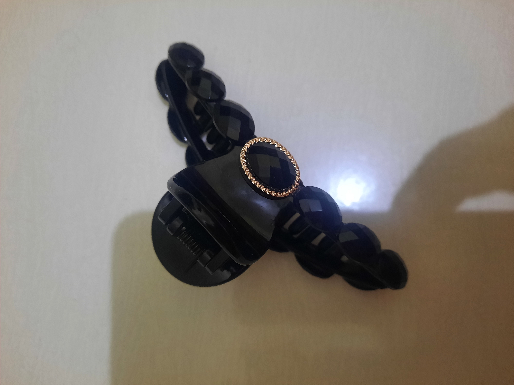 A black claw hair clip with a faceted gemstone and a gold-toned braided rim is positioned on a light-colored, textured surface, possibly a tabletop or floor. The hair clip is partially open, revealing its internal spring mechanism. The gemstone appears oval or somewhat rounded, and it is set within the gold-toned braided rim. The surface exhibits subtle shadows and highlights, suggesting the presence of ambient light, possibly from a window or overhead source. There are no people or other identifiable objects in the frame. The background is neutral and out of focus, emphasizing the hair clip as the primary subject. This image does not provide any information related to the city of Lhokseumawe, Indonesia.