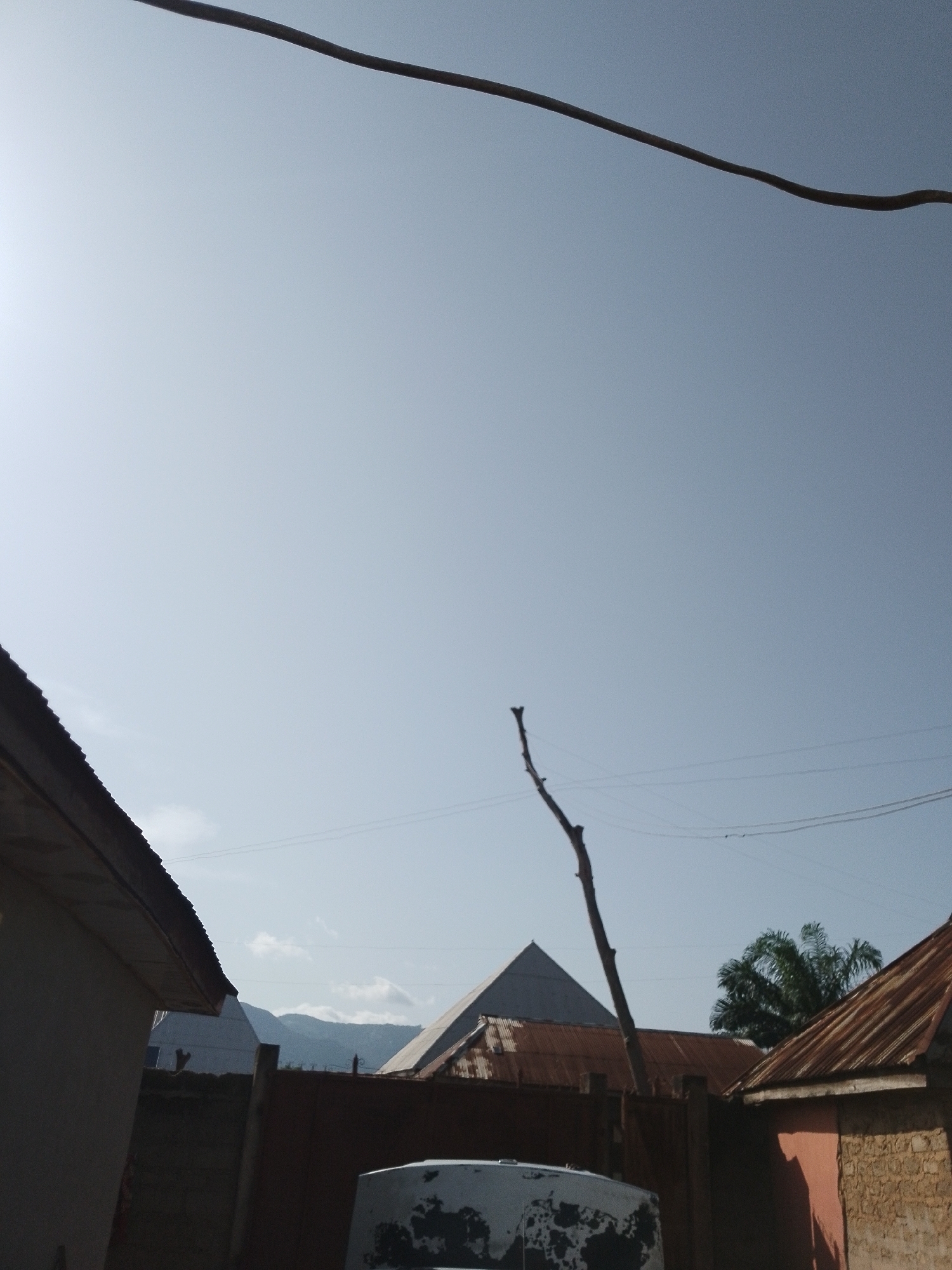 The image captures a clear sky over a residential area in Serti, Nigeria. In the foreground, a portion of a building with a dark-pitched roof is visible on the left. Behind it, a brown wooden fence separates properties. Further back, several structures with corrugated tin roofs are present, one of which appears to be a building with a pyramidal roof structure behind it. A tall, bare tree branch extends upwards into the sky. In the distance, mountains are visible under a hazy sky. Power lines are strung across the upper portion of the image. A white, partially visible object, possibly a vehicle, is in the lower right corner. No people are visible in the frame. The overall scene depicts a typical day in a rural or semi-urban setting.