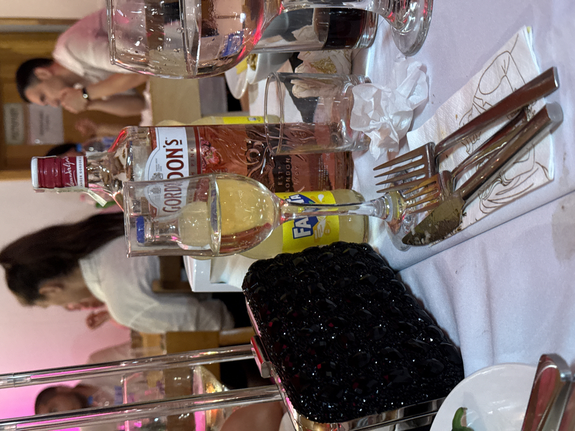 The scene is set indoors at a social gathering or restaurant in Debrashtitsa, Bulgaria, under ambient pink and white artificial lighting. The foreground features a white tablecloth with various dining and drinking items. A black, heavily beaded or sequined clutch purse with a silver clasp rests on the left. To its right, a tall wine glass contains a clear, yellowish liquid, possibly white wine. A yellow Fanta bottle is partially visible behind this glass. Centrally located is a bottle of Gordon's Pink Gin with a red cap and label depicting strawberries. Further right, an empty clear glass tumbler and a larger wine glass containing a dark liquid (possibly red wine) are present. Crinkled white paper tissues are scattered on the tablecloth. A set of silver cutlery, including forks and knives, lies on a white paper napkin adorned with line drawings of formally dressed individuals. A white plate with a green garnish is partially visible in the bottom left.
In the background, two individuals are discernible. On the left, a person with dark hair tied in a ponytail is seen from behind, wearing a white long-sleeved shirt. On the right, a man with short dark hair is observed in profile, wearing a light-colored collared shirt. He is seated, with his hands clasped under his chin, indicating a contemplative or attentive posture. The background features light-colored walls and what appears to be a doorframe, with other blurry figures and furniture suggesting a busy dining environment.