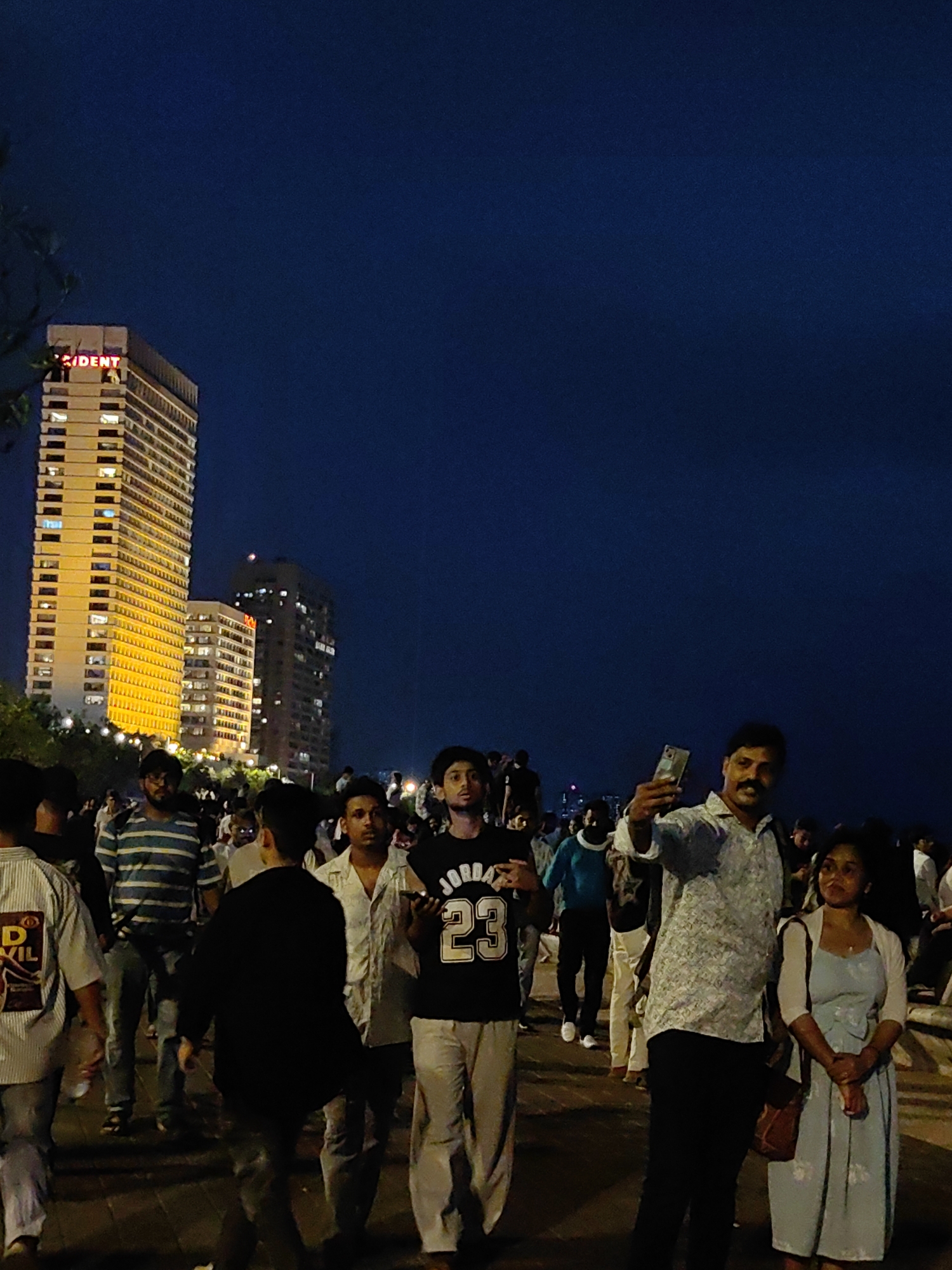 This is a photograph taken at night in Mumbai, India, likely along the Marine Drive promenade. The foreground shows a crowd of people walking and interacting. One man in the center is taking a selfie with his phone, while a woman stands beside him. The background features illuminated high-rise buildings, including a prominent one with "PRESIDENT" lit up in red letters at the top. The sky is a deep, dark blue, suggesting it is late evening or night. The ground appears to be paved, characteristic of a promenade.