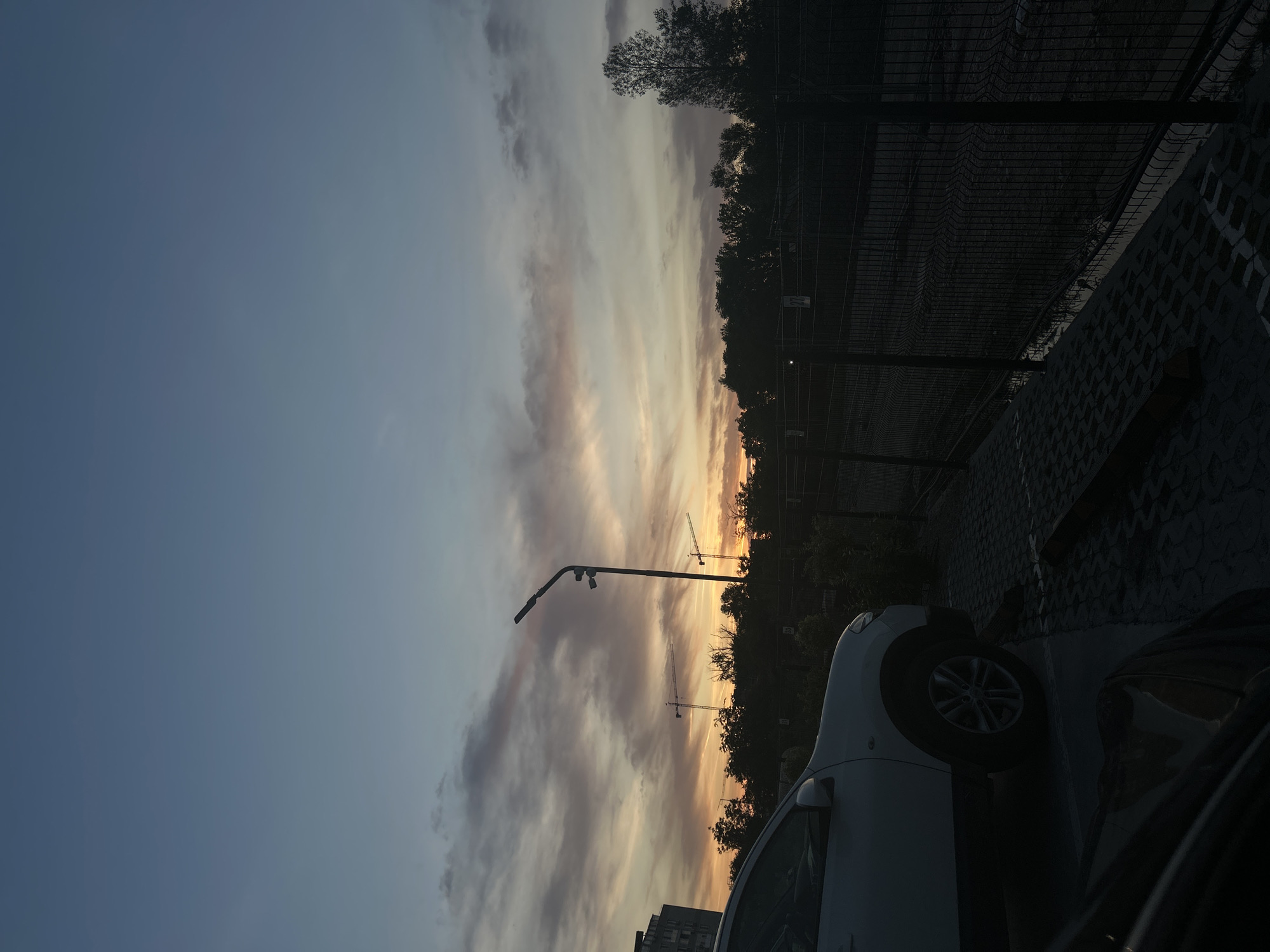 The image shows a parking lot at sunset in Plovdiv, Bulgaria. The sky is visible with clouds illuminated by the setting sun, showing orange and pink hues. There are two construction cranes in the distance silhouetted against the sky. A street lamp stands next to a white car parked in the lot. A dark-colored car is also partially visible. A chain-link fence borders the parking lot. Trees are visible in the background. A building can be seen in the lower-left corner of the image.