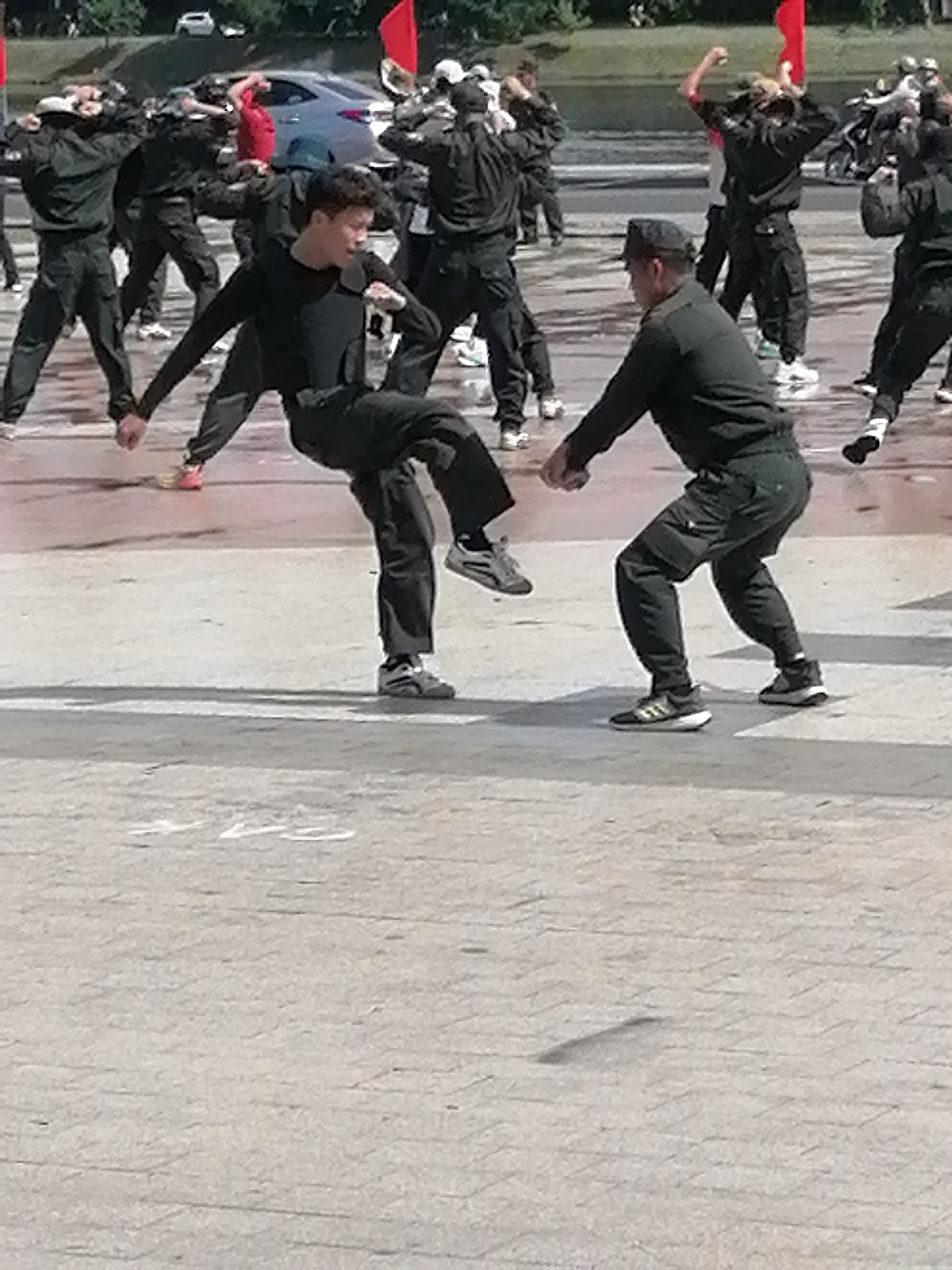 The image depicts a group of individuals, appearing to be young men, engaged in what seems to be a synchronized martial arts or dance practice outdoors. The individuals are dressed in dark, loose-fitting clothing, some wearing military-style caps. The central action focuses on two individuals in the foreground. One person, dressed in black, is in mid-air, executing a high kick with their right leg extended. The other individual, also in dark attire and wearing a cap, is positioned in front of them, facing away from the viewer and appearing to be reacting to or anticipating the kick. The background shows other individuals in similar poses, suggesting a larger group activity. The environment appears to be an open, paved area, possibly a public square or training ground. In the background, there are hints of vehicles and trees, suggesting an urban setting. The lighting indicates it is daytime. The specific location in Da Lat, Vietnam, cannot be determined from the visual information provided in the image.