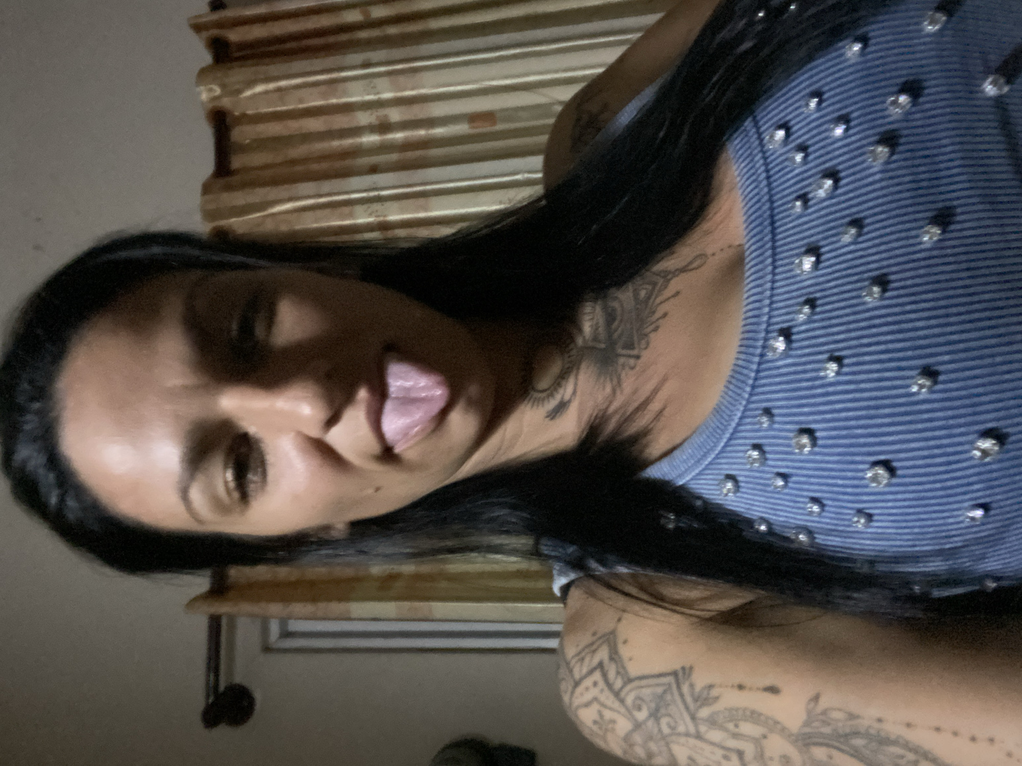 A woman with dark hair and light skin, adorned with tattoos on her neck and arm, poses indoors with her tongue sticking out. She is wearing a blue striped top with shimmering embellishments. Behind her, there is a window covered with striped curtains. The location is potentially in Mũi Né, Vietnam. Additional details are limited without further context.