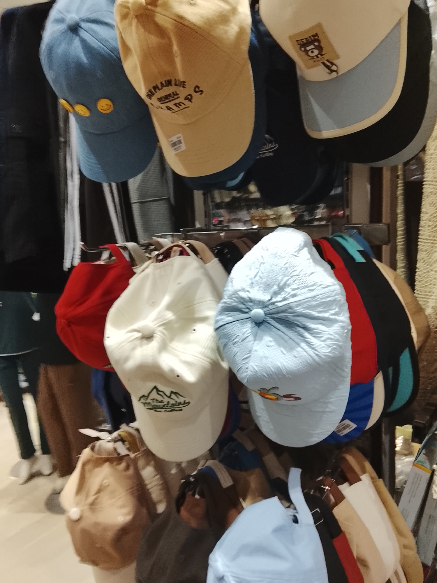 The image shows a rack of baseball caps in a retail environment, likely in Gulgasht Colony, Pakistan. The caps are arranged on hangers, with several colors and styles visible.
The top row of caps includes blue, tan, and light blue options. The blue cap has smiley face icons. The tan cap has the words "The plain live General Camps" on it. The light blue cap has a patch on it.
Below, there are caps in red, white, and blue. The white cap has a mountain logo on it. The blue cap has an image on it.