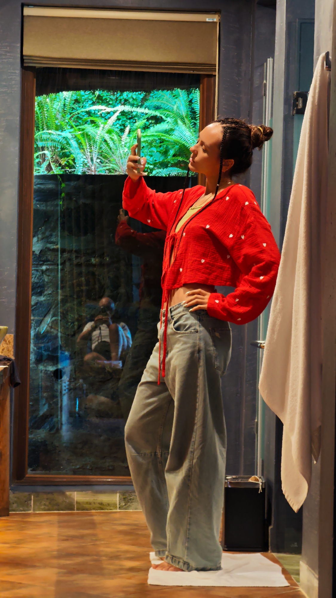 An indoor scene in Mu Cang Chai, Vietnam, depicts a female subject taking a selfie with a smartphone in front of a large window. The adult subject, featuring braided dark hair styled into a high bun, wears a red long-sleeved cropped top adorned with white heart patterns, a light green undergarment, and light blue baggy jeans. She stands on white rectangular foot covers positioned on a white towel. Her right hand holds a green-gold smartphone at eye level, directed towards herself, while her left hand rests on her hip. The large window has a dark wooden frame and a partially visible beige roller blind at the top, revealing vibrant green foliage outside. The window's lower section reflects a blurred individual, likely the photographer, wearing a white t-shirt and shorts, holding a camera. The interior walls are dark blue-grey and textured. A white towel hangs on a metal rack on the right. A small black trash can is visible in the lower right corner. The floor is brown with a subtle pattern.