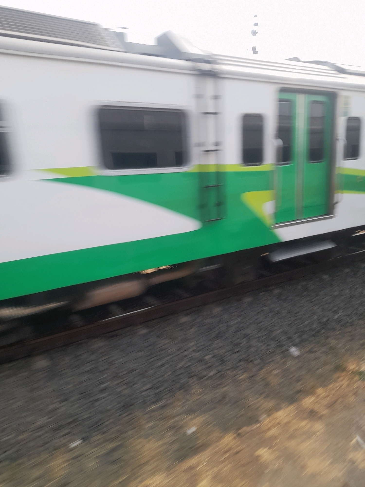 The image shows a train moving on a railway track. The train is white with a green and yellow stripe along its side. The windows of the train are dark and appear to be tinted. The train doors are green. The train is captured in motion, resulting in a blurred effect. The railway track is visible in the foreground, with gravel and dirt surrounding it. In the background, there are some blurred structures that could be buildings or utility poles. The sky is overcast or hazy. The image appears to have been taken from another moving vehicle or from within the train itself, giving a dynamic perspective of the train's movement. The image is a photograph of a train at the Mojokerto railway station in Mojokerto, Indonesia.