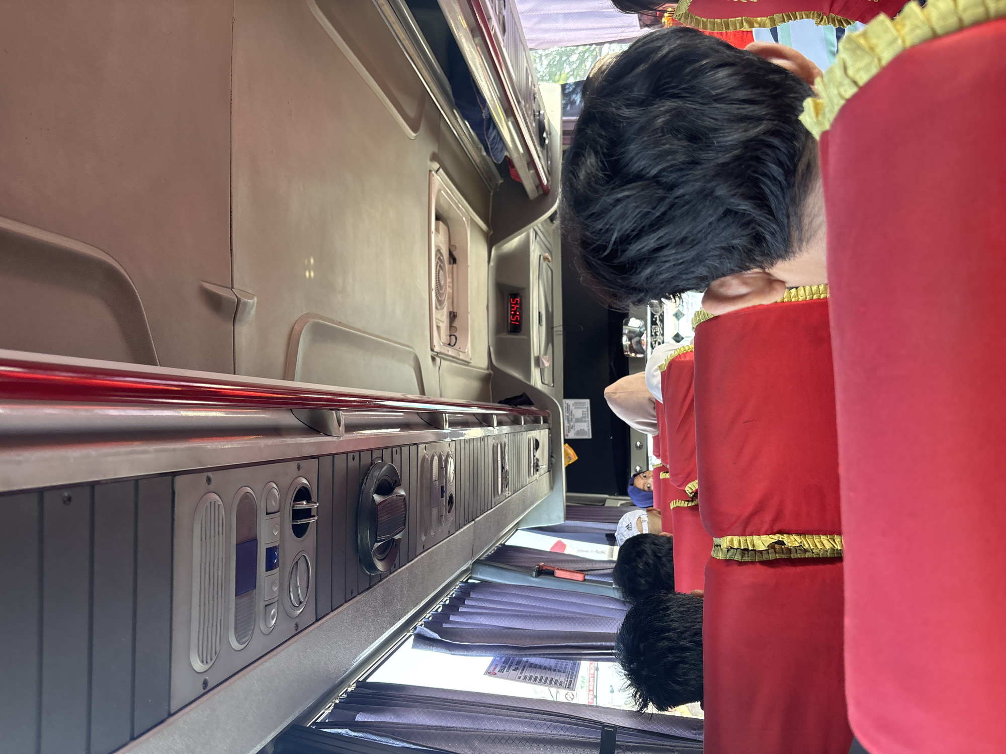 The image shows the interior of a bus, likely in Cikampek, Indonesia, with passengers seated. The viewpoint is from behind a passenger in the foreground, looking towards the front of the bus. Red seats with gold trim are visible. The ceiling of the bus features overhead compartments, air vents, and a digital clock displaying "15:45". Outside the window, purple curtains are drawn, and a sign is partially visible. The overall lighting suggests it is daytime.