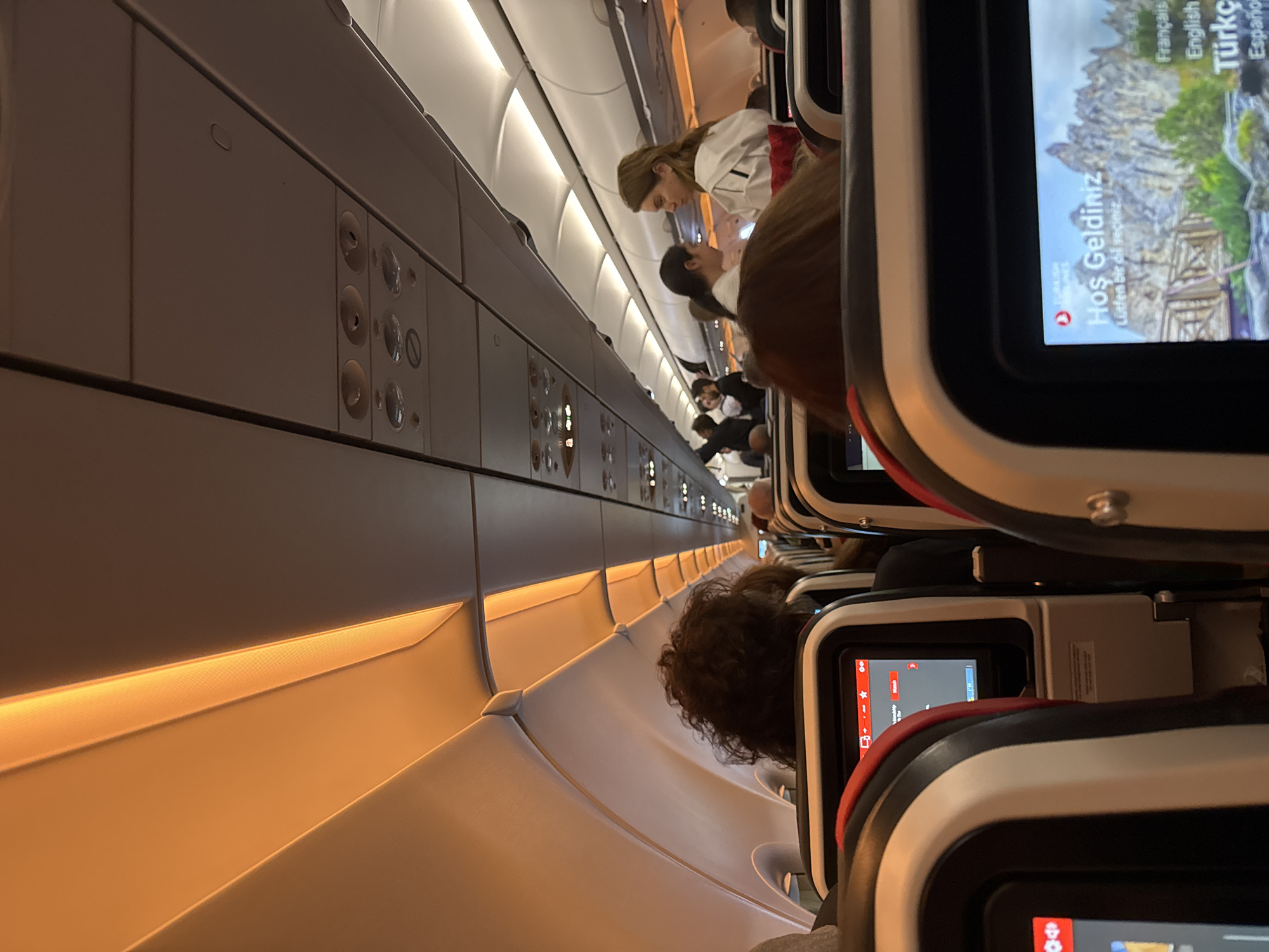 The image captures an interior shot of an airplane cabin, showing several rows of seats occupied by passengers. A flight attendant is visible interacting with a seated passenger. The aircraft features overhead compartments along the upper section of the frame, with air vents and reading light controls. Each seat is equipped with a personal entertainment screen, one displaying a welcome message in multiple languages, including Turkish. The image does not provide any information to ascertain the location in the city of Geghanist, Armenia.