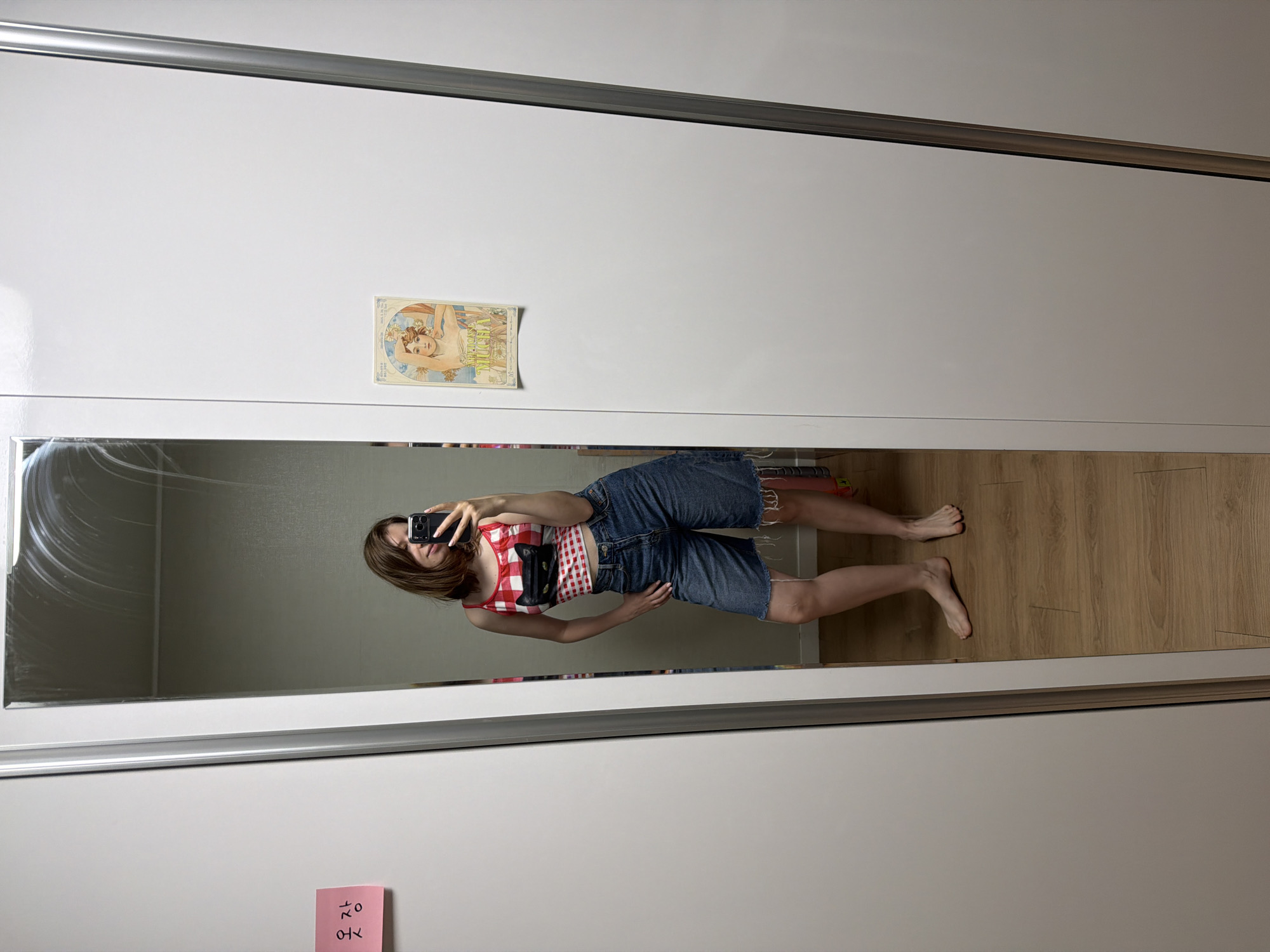 A woman takes a mirror selfie in a room that appears to be a closet or dressing area. She is wearing a red and white checkered tank top with a black cat appliqué on the chest and denim shorts with frayed hems. Her left hand is on her hip, and she is holding her phone in her right hand to take the picture. A pink sticky note with Korean writing is affixed to the closet door to her left. To her right, on the same closet door, is a decorative print of a woman in a flowing gown. The floor is wood-paneled.