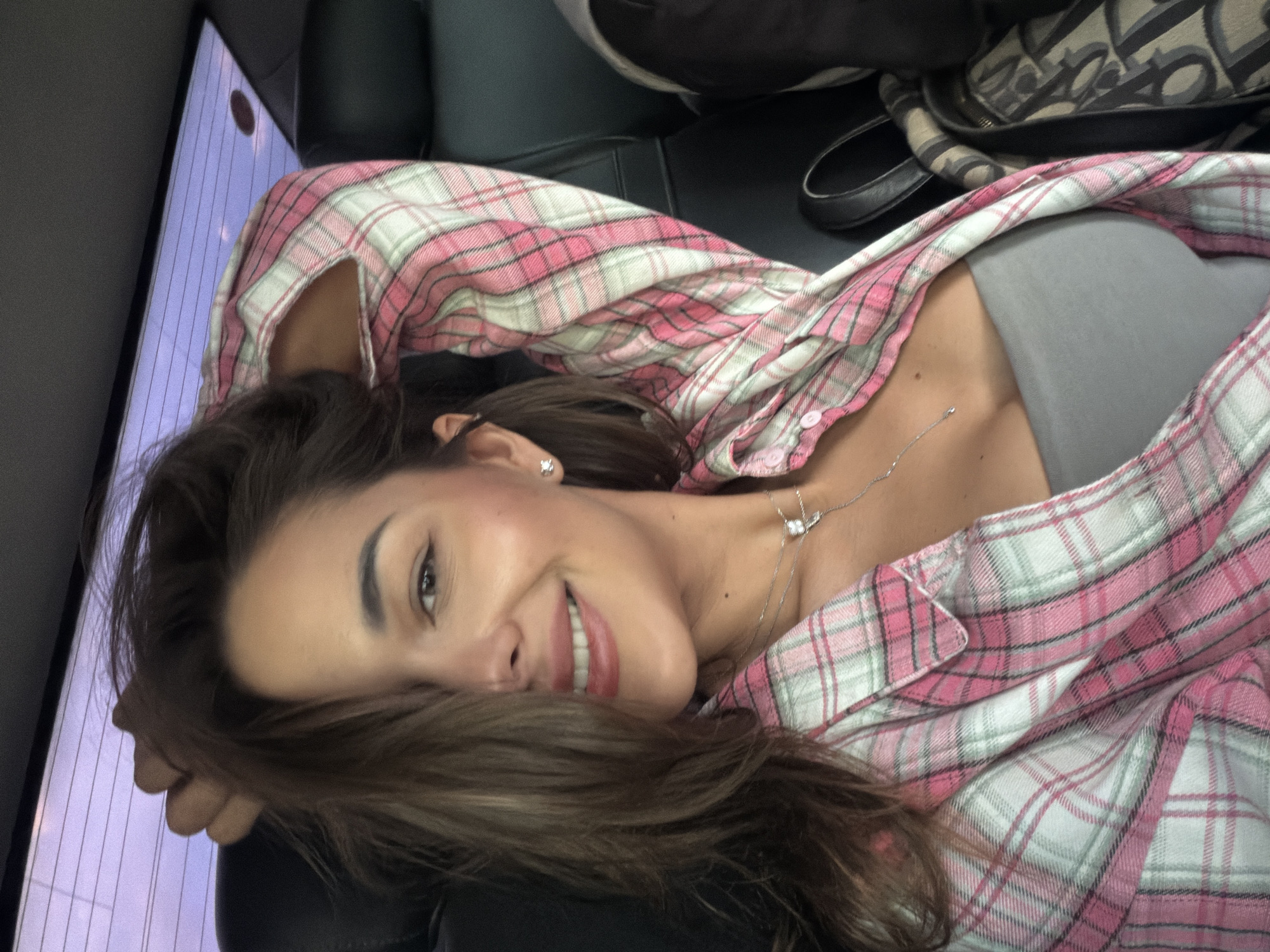 A young woman is lying down, smiling at the camera. She is wearing a pink and white plaid shirt over a grey top and a delicate necklace. Her hair is brown, and she has a small earring. She appears to be lying on a black leather surface, possibly a car seat. In the background, there's a screen and a patterned bag. The location is not discernable from the image. There is no information available about the city of Yemelyanovo, Russia.