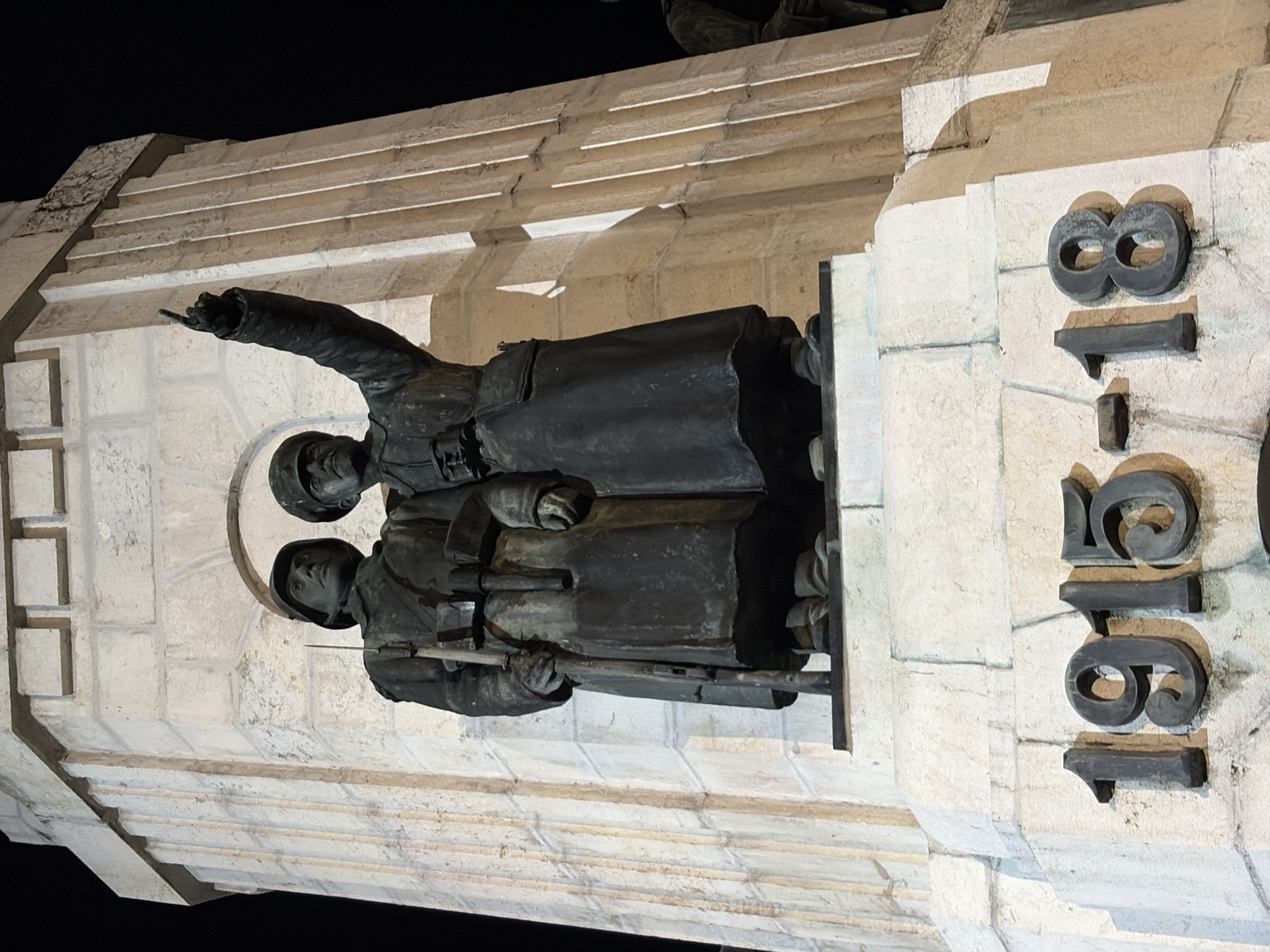 The image depicts a stone monument featuring two bronze soldier statues. The soldiers are wearing helmets and carrying rifles. One soldier is pointing. To the right of the statues, the numbers "1918" are inscribed vertically in large, raised characters on the stone. The monument appears to be located in an outdoor setting, illuminated by artificial light. The specific location is likely Veliko Tarnovo, Bulgaria.