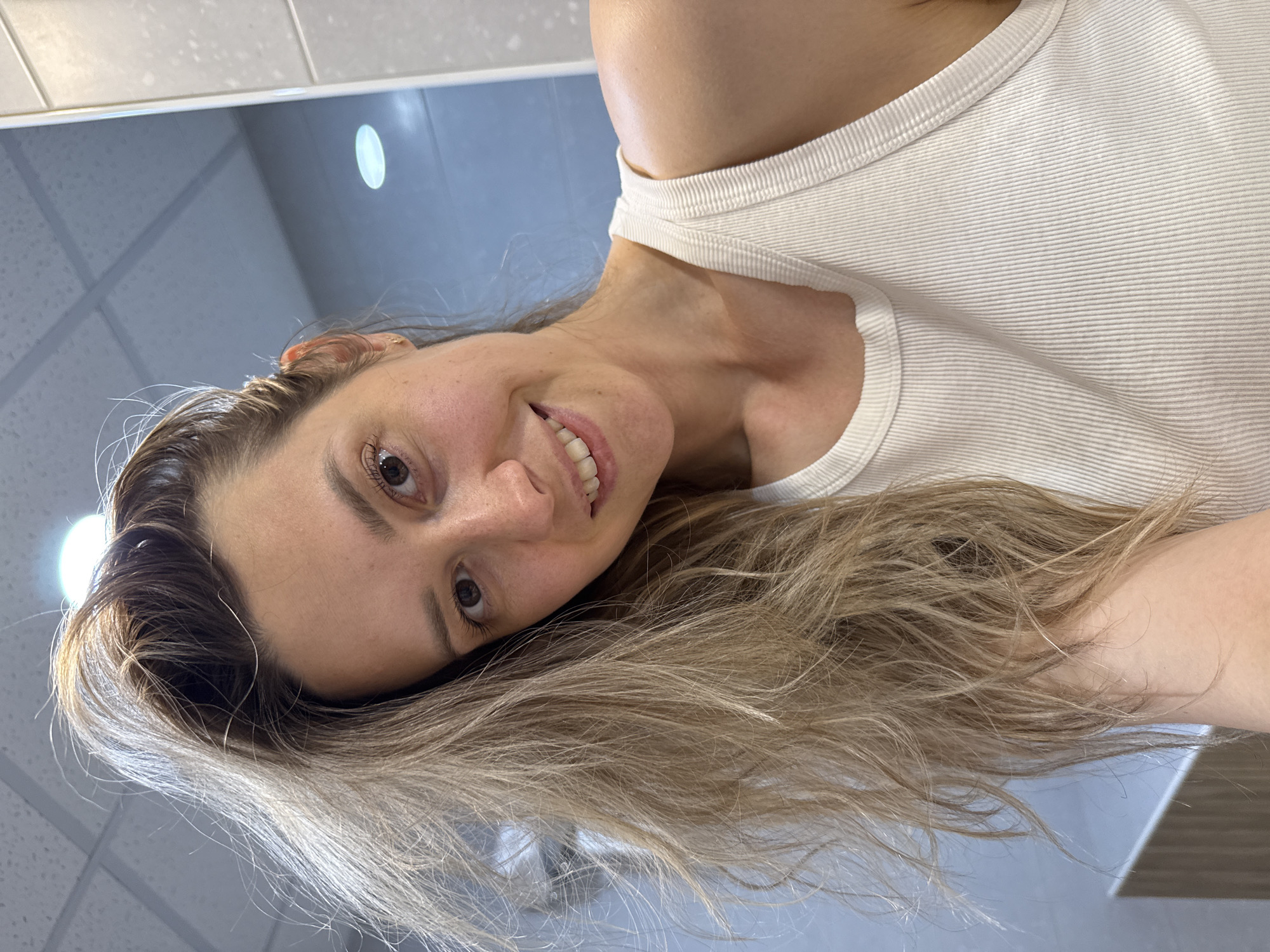 A female subject is captured in a close-up selfie, smiling subtly and looking directly at the camera. She has long, light brown hair, appearing somewhat disheveled, with strands falling over her left shoulder and chest, while some are pulled back loosely on the right. Freckles are visible across her nose and cheeks, and she has brown eyes. She wears a white, ribbed sleeveless tank top and a small, gold-colored stud earring in her right earlobe. The background is an indoor setting, characterized by light-colored tiled walls on the right and a lighter blue-grey wall section further left. A white grid ceiling with square panels and visible recessed lighting fixtures suggests a public or commercial restroom or changing area. A bright circular light source is reflected in the upper right background.