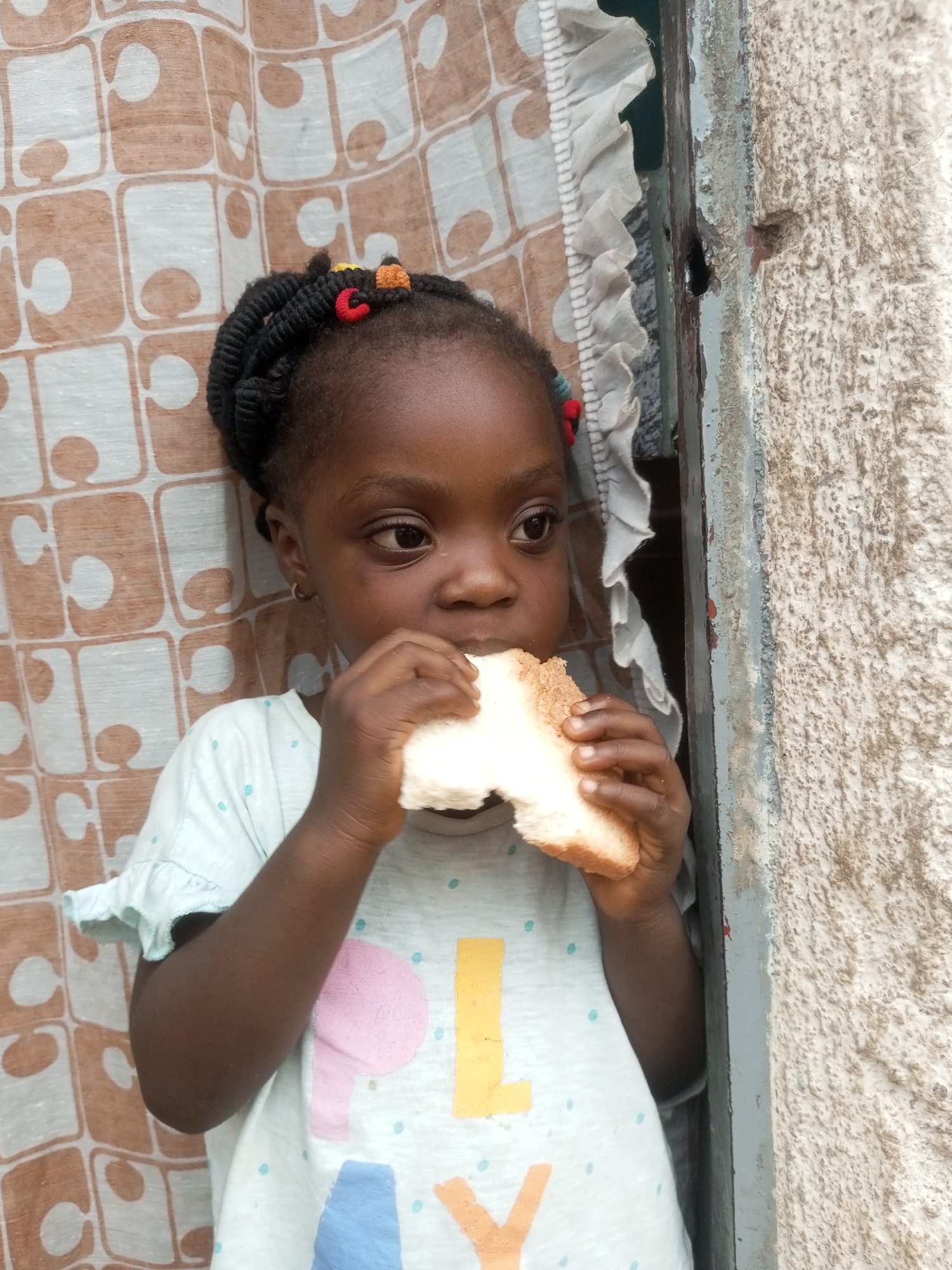In this close-up, waist-up shot, a young Black girl is captured in an indoor setting, likely a home, as she eats a piece of white bread. She is positioned to the left of the frame, leaning against a patterned curtain with a brown and white geometric design. The curtain has a frilled edge on its right side. To the right of the girl is a weathered, light gray concrete wall and a dark-colored door frame, suggesting an urban environment. The girl has dark, braided hair adorned with small red, yellow, and orange beads. She is wearing a light blue t-shirt with a repeating pattern of light blue dots and colorful graphic elements, including what appear to be letters or shapes in pink, yellow, and blue. She holds a piece of bread in both hands, bringing it to her mouth to take a bite. Her gaze is directed to her right, away from the camera. The lighting is somewhat diffused, with shadows subtly defining the contours of her face and hands. The scene conveys a moment of everyday life, focusing on the child's simple act of eating.