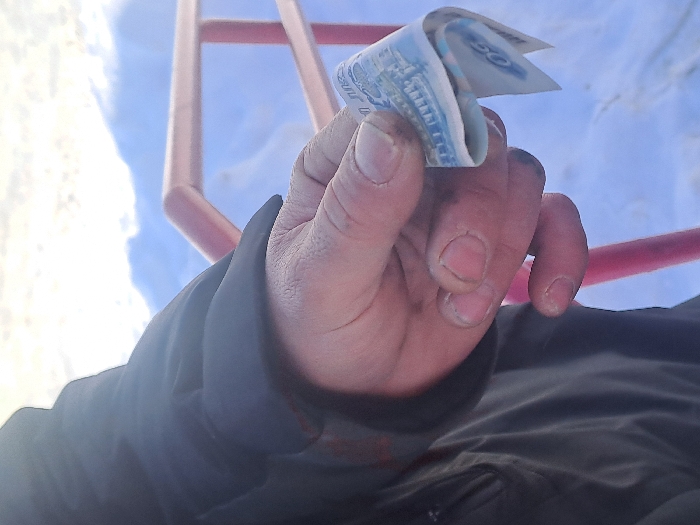 A close-up, outdoor shot in Bolshaya Elhovka, Russia, depicts an adult right hand holding a folded banknote. The hand, exhibiting visible dirt or grease on the fingertips and unmanicured nails, is partially covered by a dark, possibly winter jacket sleeve. The banknote, identified as a light blue and white Russian 50 Ruble note, is held between the thumb and index finger. In the mid-ground, a pink/red metal railing or pipe is present. The background is dominated by snow-covered ground, indicating a winter environment under bright, possibly overcast, daylight conditions.