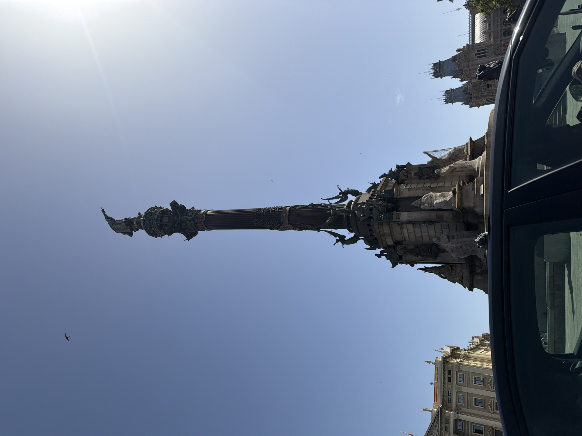 A low-angle, full shot captures the Monument to Columbus in Barcelona, Spain, under a clear blue sky. The monument, a tall, dark column topped with a statue of Christopher Columbus, is detailed with several ornate sculptures and figures at its base. To the left of the monument, a grand, light-colored building with multiple windows and ornate architectural details stands. To the right, another building with distinctive gray spires can be seen. In the foreground, the dark, reflective surface of a car is visible, obscuring the lower part of the scene. A single bird is seen flying in the vast expanse of the sky. The sun is shining brightly, casting a lens flare in the upper right corner of the image. The overall scene depicts a notable landmark and its surrounding architecture on a clear, sunny day.
