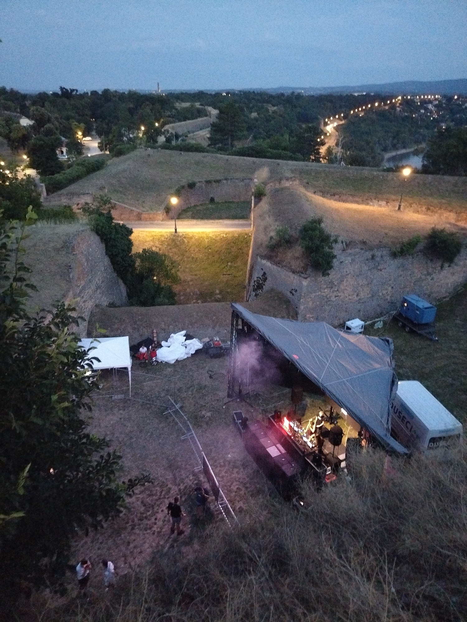The image captures an aerial view of an outdoor event setup at dusk, possibly at the Petrovaradin Fortress in Petrovaradin, Serbia. A large, angled stage covered by a gray tarp is set up in the foreground, with lights illuminating the performers on it. A white van with the word "DUSECI" visible on its side is parked to the right of the stage. In the background, a blue container on a trailer sits near the stage. Further back, a white tent stands, with several people gathered around it. The surrounding environment consists of grassy hillsides and what appear to be the fortified walls of the fortress, with scattered trees and lights from the distant city visible on the horizon. Several individuals are scattered throughout the scene, some observing the stage and others moving around the site. The overall atmosphere suggests a concert or festival preparation in progress.