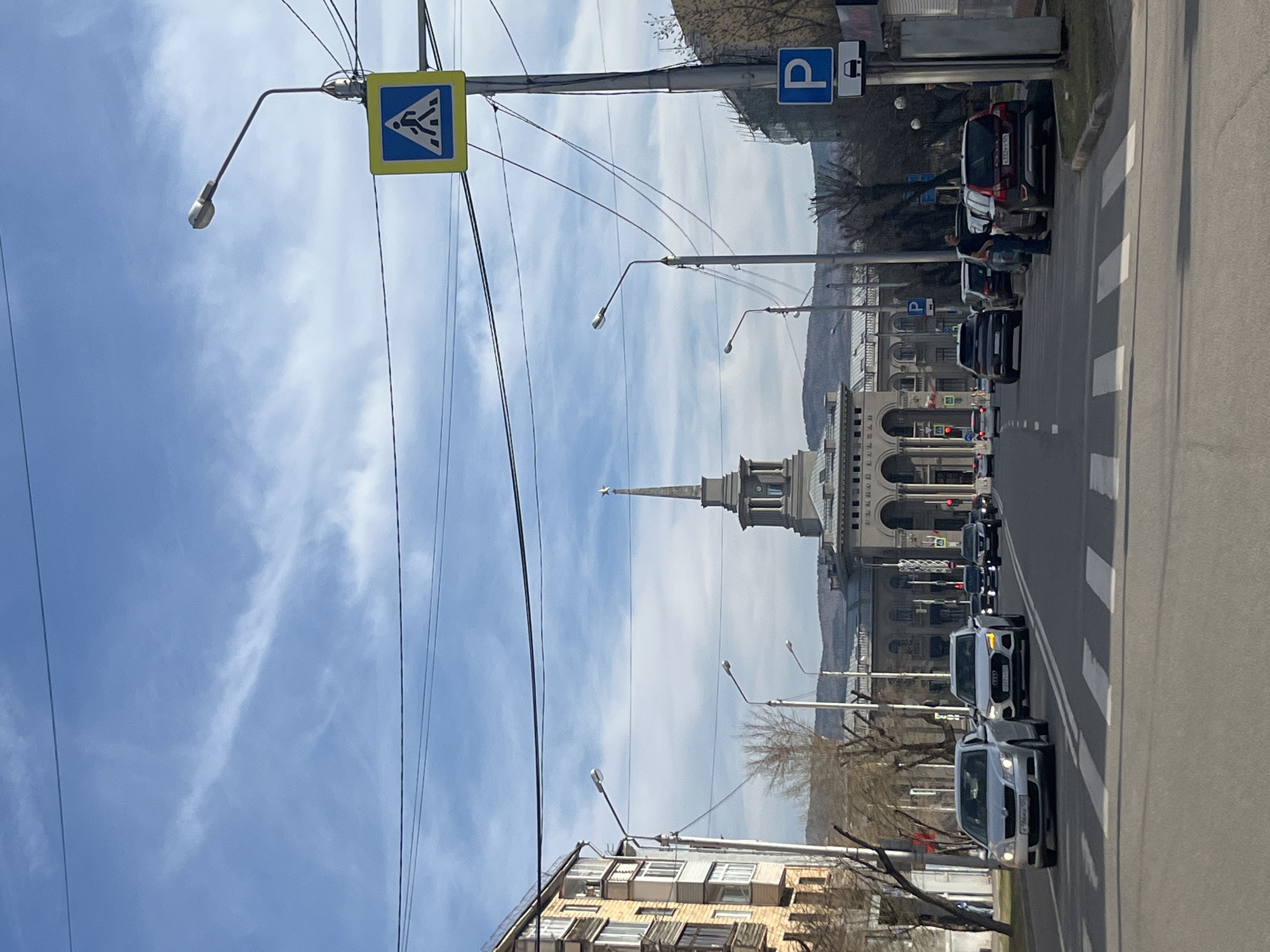 The image shows a street scene in Krasnoyarsk, Russia, with a view towards the city center. The sky is partly cloudy with thin, scattered clouds. The street is lined with buildings on both sides. A prominent building with a spire and arched facade is visible in the distance, likely a government or administrative building. The street has several parked cars on the right side, and more cars are driving in the street, with visible traffic lights in the background. There are lampposts along the street and power lines overhead. A pedestrian crossing sign is visible on the left. A person is standing next to a parked SUV near the sidewalk. The buildings along the street are mostly residential. Trees are visible in the background and along the street, with bare branches, suggesting it's early spring.