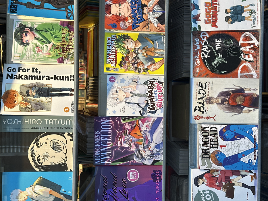 The image displays a detailed view of a retail manga book display across three horizontal shelves. Approximately fifteen distinct manga volumes are fully or partially visible, arranged vertically with their covers facing outwards.
On the top shelf, from left to right, visible titles include a blue-haired male character with a backpack; "YOSHIHIRO TATSUMI ABANDON THE OLD IN TOKYO" featuring a distressed face; "Go For It, Nakamura-kun!! Volume 2" depicting two male characters; and "The Apothecary Diaries" with a female character in green attire.
The middle shelf features "Second Hand Love" (partially visible); "NEON GENESIS EVANGELION" showcasing an Eva unit and pilot; "WAKABA WON'T GIVE UP! Volume 1" with a white-haired female character; "MY HERO ACADEMIA" with multiple smiling characters; and "PHANTOM BUSTER" with male characters.
The bottom shelf presents "DRAGON HEAD 1" with two blue-skinned figures; "BLADE OF THE IMMORTAL OMNIBUS 1" featuring a samurai and a female in red; "THE GIRL WHO RAISED THE DEAD" with a skeletal hand motif; and "GOGO MONSTER" depicting two young male characters in school uniforms.
The books are housed on dark-colored, likely metal, shelving units, some with thin vertical dividers. Below the main display rows, numerous additional book spines are visible, indicating deeper shelving or stacks of inventory. The scene is brightly lit by an unseen source, causing minor reflections on some covers. The environment is an interior retail setting, specifically a bookstore or comic shop specializing in Japanese manga.
This scene is located within a bookstore or similar retail establishment in London, United Kingdom. The precise street address or specific store identity is not discernible from the provided image.