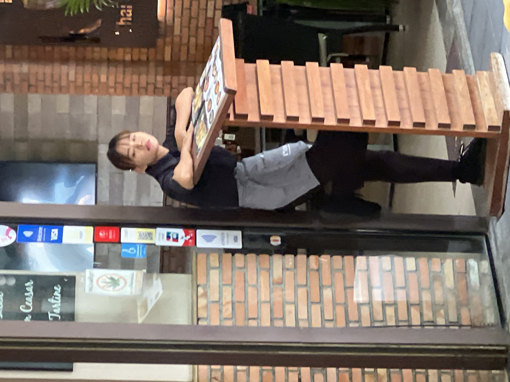 A young woman, appearing to be a server, stands behind a wooden menu stand at what seems to be an establishment in the city of Ban Bang Rak, Thailand. She is wearing a black short-sleeved shirt, a light gray apron, and black pants. Her arms are resting on the top of the menu stand, and she is looking slightly upwards and to her left. The menu stand displays several pages with images of food items. To the left of the woman, a brick wall is visible, and to the far left, a glass door with signage is present. On the door, various stickers are affixed, including a marijuana leaf symbol with a prohibition sign. The background shows more brickwork, a dark sign with the word "thai" partially visible, and some seating areas. The overall lighting suggests it might be daytime or early evening.