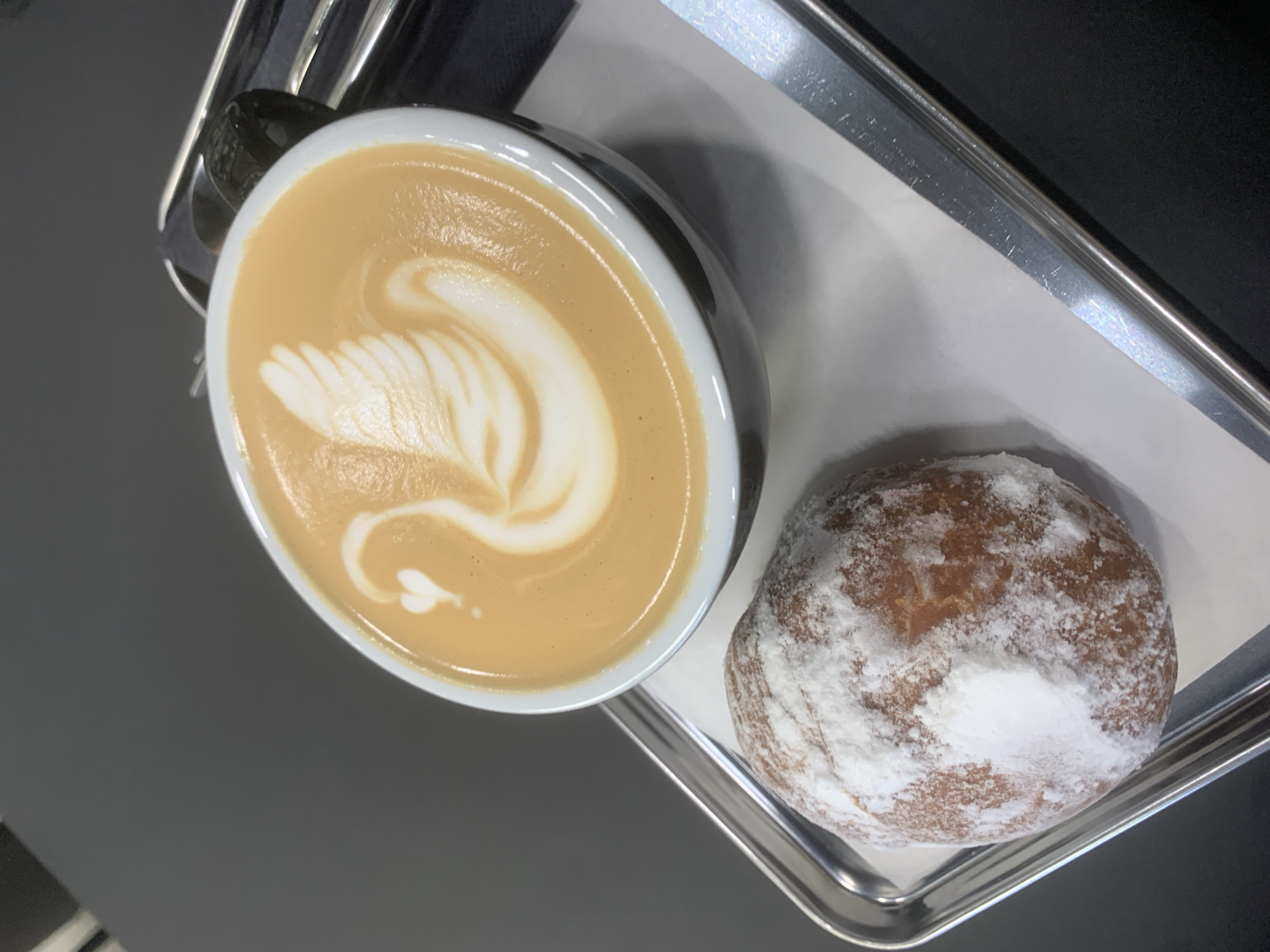 A top-down shot shows a latte and a powdered donut on a silver tray. The latte, in a black mug with a white interior, features latte art in the shape of a swan. The round donut is heavily dusted with powdered sugar. The tray is lined with white parchment paper and is placed on a dark gray surface. No people are visible. The location is unknown, and therefore cannot be determined to be Izhevsk, Russia.