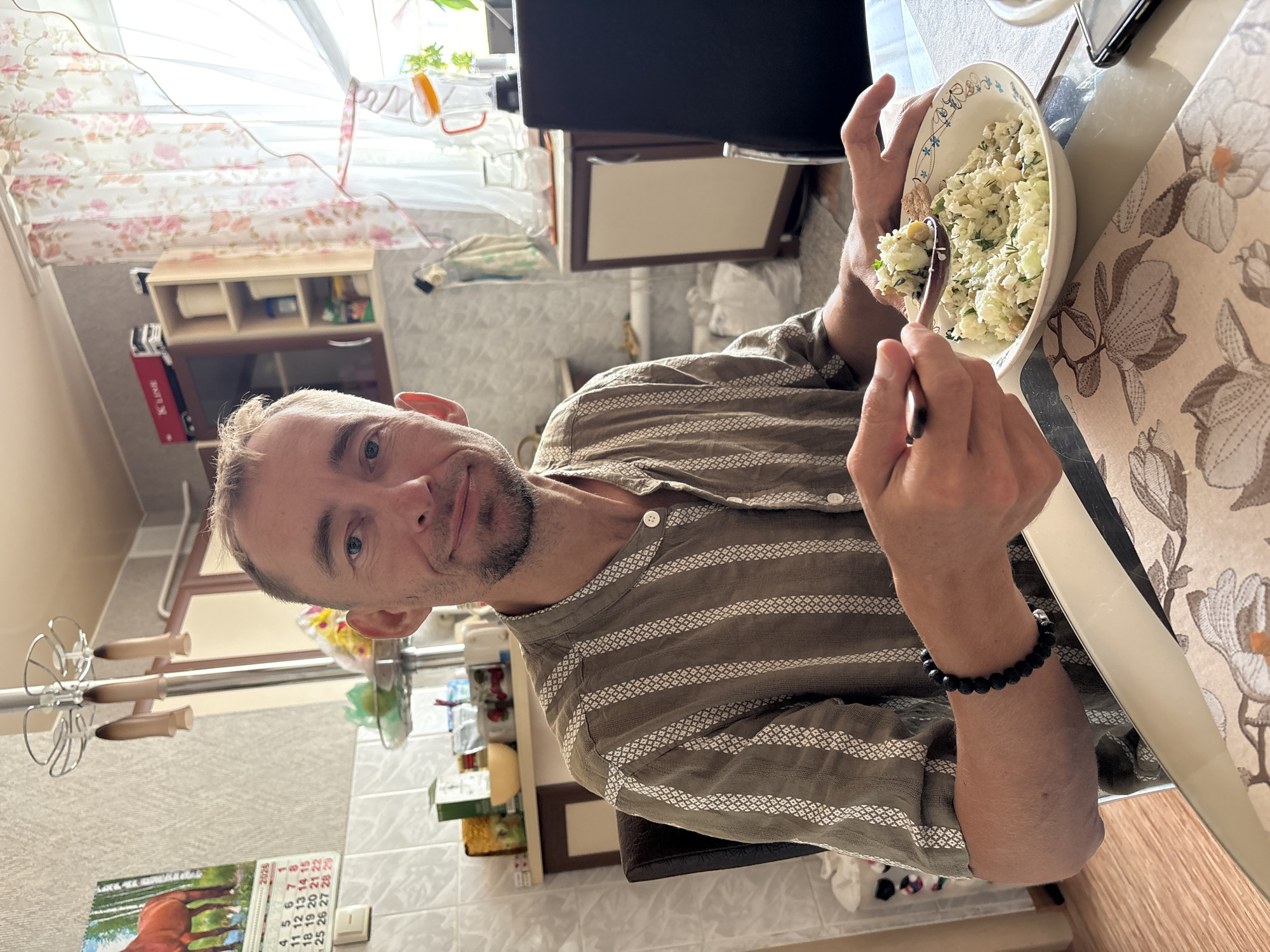 A male individual is depicted seated at a dining table in what appears to be a residential kitchen in Ulyanovsk, Russia. The man, with short, light-brown hair and a beard, is wearing a striped, collared shirt in a khaki-brown hue with white patterned stripes and a black beaded bracelet on his left wrist. He is smiling faintly and looking directly at the camera while holding a wooden spoon in his right hand, poised to eat from a white bowl containing a rice or grain-based salad mixed with green herbs and legumes. A floral-patterned placemat in beige and brown tones is beneath the bowl, and a dark-colored smartphone rests on the table to the right.
The background shows a brightly lit interior. To the left, a white tiled wall transitions to textured light-colored wallpaper. A 2025 calendar featuring a horse in a pastoral setting is visible on the wall, specifically showing April. A metal hanging rack with three beige items is mounted above shelves containing various kitchen items, including a yellow bowl and packaged goods. To the right, a window adorned with sheer, white curtains featuring a pink floral pattern allows strong natural light into the room. A blender with an orange lid, a clear glass pitcher, and other kitchen furniture, including a light-colored cabinet, are visible near the window.