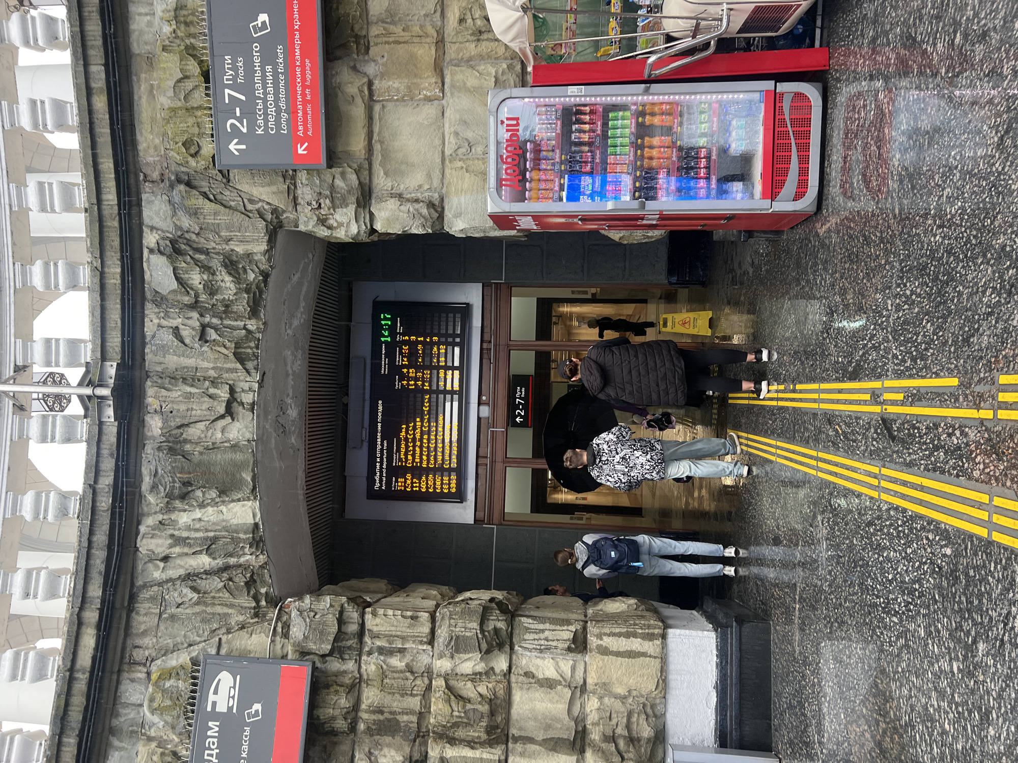 The image shows the entrance to a train station in Sochi, Russia. The entrance is flanked by stone walls and a sign pointing to tracks 2-7 and long-distance tickets. A vending machine stands to the right of the entrance. A train schedule is displayed above the entrance. Several people are entering or exiting the station. The floor is wet and reflecting light. Yellow tactile paving guides visually impaired passengers. A sign on the lower left reads in Russian "to the platforms, ticket offices".