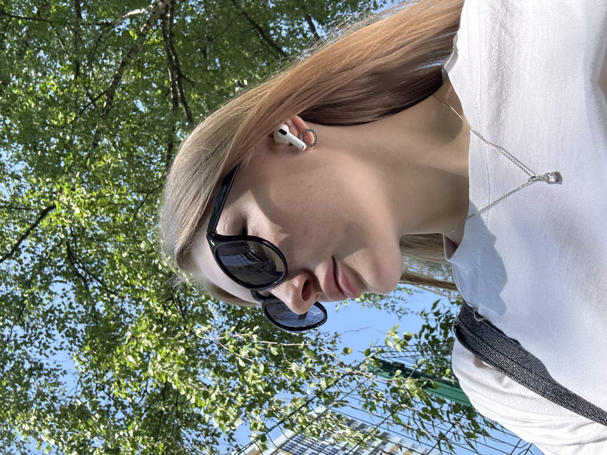 A low-angle, upward-facing shot depicts a young woman with blonde hair, wearing sunglasses, an earbud in her left ear, and a white t-shirt. A silver necklace with a diamond pendant adorns her neck.
The background consists of green foliage and a blue sky, with glimpses of a chain-link fence and urban structures, suggesting an outdoor setting in Perm, Russia.