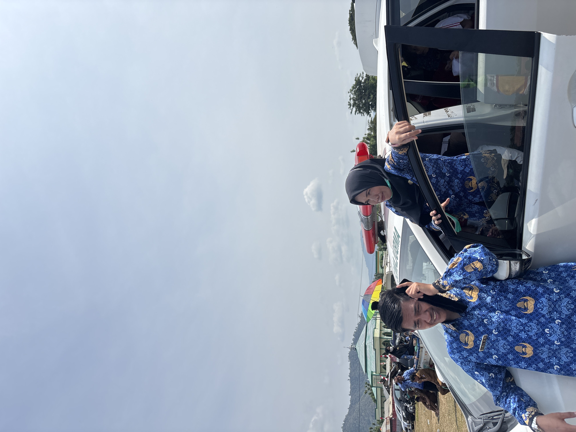 In Torosiaje, Indonesia, under a vast, pale blue sky with scattered clouds, a group of people are gathered outdoors. In the foreground, a man in a blue patterned shirt with gold embellishments leans against a white vehicle, holding a black object to his ear, appearing to be on a phone call. To his right, a woman wearing a black hijab and a matching blue patterned outfit leans out of the open passenger side door of the same vehicle, her left hand resting on the car's frame. The vehicle has a red flashing light on its roof.
In the background, several other vehicles are parked, and a colorful umbrella stands out. A building with a green roof and white walls, possibly a mosque, is visible in the distance, set against a backdrop of rolling hills. Several individuals are seen in the mid-ground, some sitting on the ground and others standing near the vehicles. The overall scene suggests an outdoor gathering or event taking place in a semi-rural or suburban area.