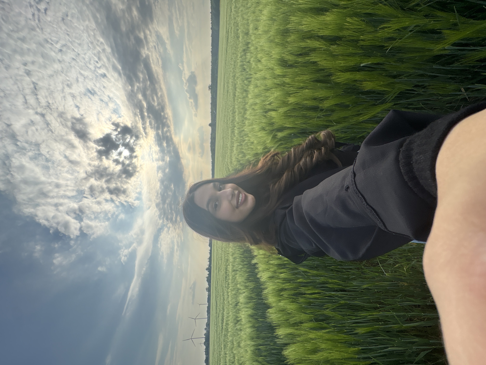 A woman is posing for a selfie in a field of green crops. She is wearing a black jacket. The sky is cloudy, and a few wind turbines are visible in the background. Based on the presence of wind turbines and agricultural fields, the image is consistent with the rural landscape around Wegberg, Germany.