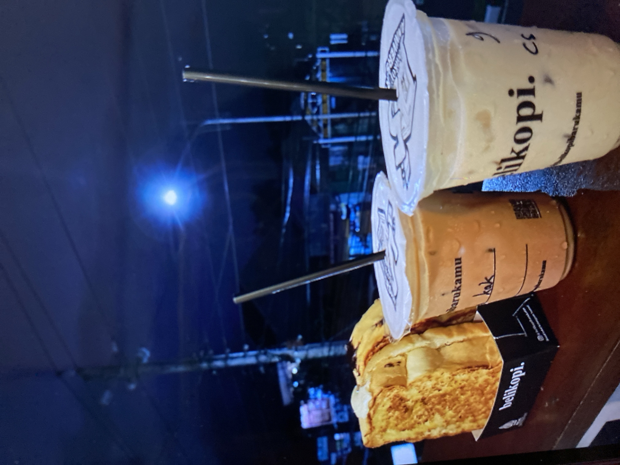 In Kalitinggar, Indonesia, on a dark, possibly nighttime, street, two iced beverages and a portion of french toast are displayed on a wooden surface. The beverages are in clear plastic cups with white lids and black straws. The cup on the left contains a caramel-colored drink, while the cup on the right holds a lighter, creamy beverage. Both cups have "belikopi." printed on them. The french toast is golden brown and appears to be topped with a dollop of white cream and drizzled with a dark sauce, possibly chocolate. It is presented in a black paper container also bearing the "belikopi." logo. In the background, out-of-focus lights and structures suggest a street scene with buildings and possibly a lamppost emitting a bright blueish light. The overall ambiance is casual and indicative of a nighttime cafe setting.