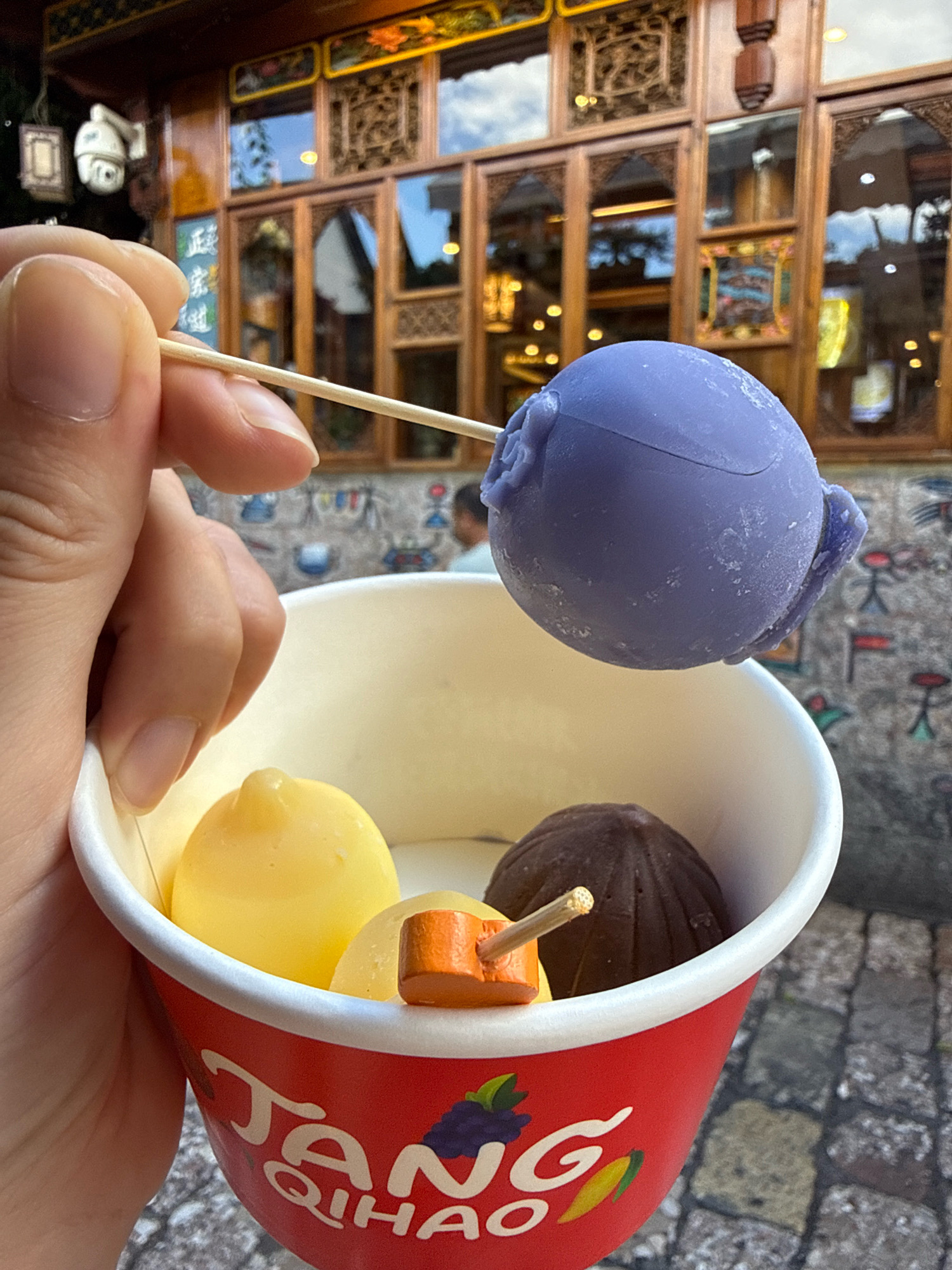 In Gucheng District, China, a hand holds a wooden skewer with a purple, rose-shaped ice cream scoop poised above a red paper cup filled with various ice cream flavors. The cup has white text and colorful fruit graphics. Inside the cup are three ice cream spheres: a yellow one, a dark brown one, and an orange piece with a skewer. The background features traditional wooden architecture with ornate carvings and reflective windows, suggesting a historic or culturally significant area. A security camera is visible on the left side of the building. The ground is paved with cobblestones.