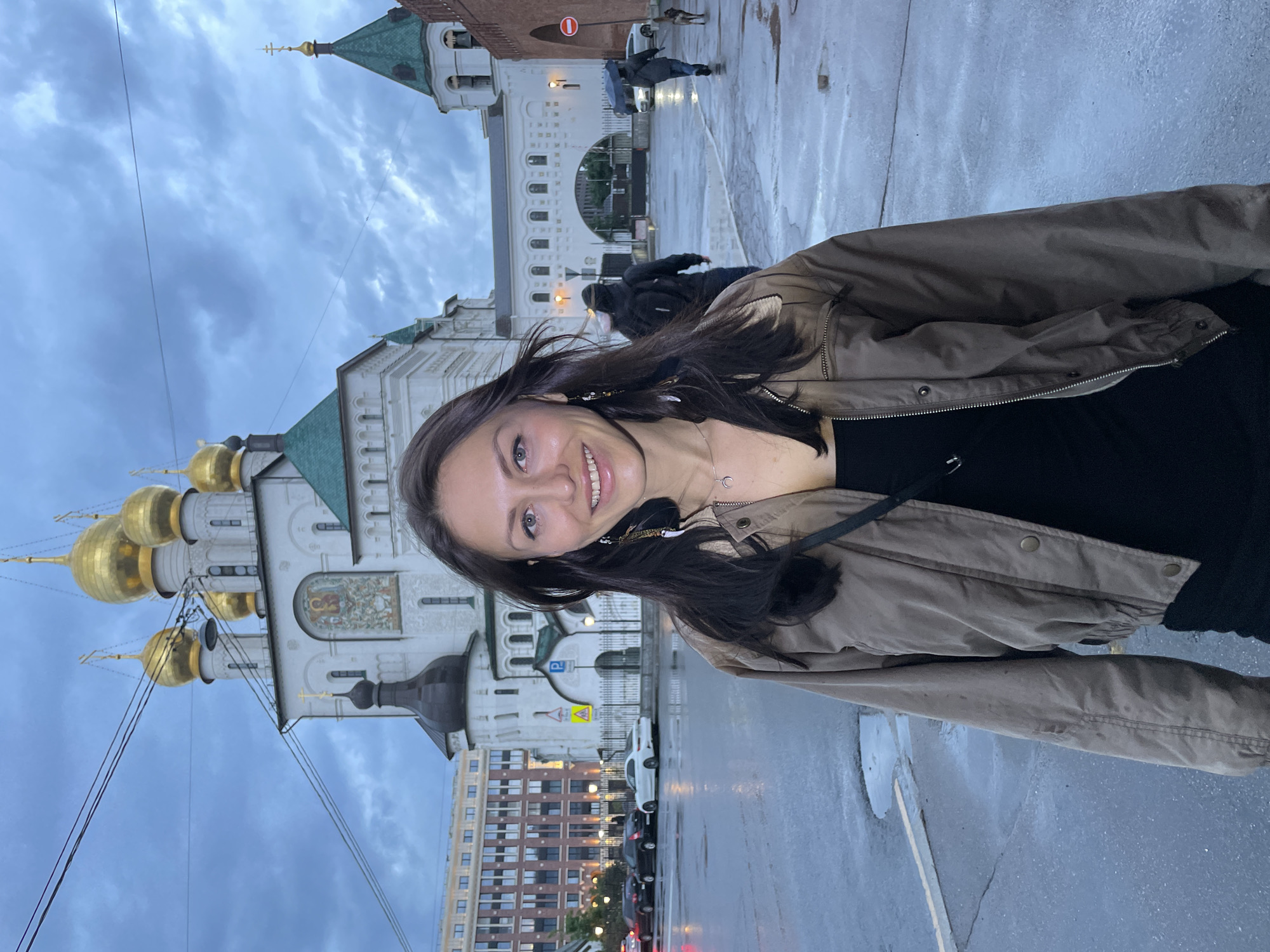 A woman with dark hair and blue eyes is standing in the foreground of the image, smiling at the camera. She is wearing a brown jacket over a black top and a silver necklace. Her earrings are long and feathery. Behind her, a large white building with golden domes stands under a cloudy sky. The ground is wet, indicating recent rain, and there are reflections on the pavement. Several cars are parked on the street, and other people are visible in the background, some with umbrellas, suggesting it is raining or has recently rained. The architecture of the building with golden domes is characteristic of Russian Orthodox churches. Given the presence of such a building and the overall aesthetic, this location is likely a religious site in Saint Petersburg, Russia, possibly a cathedral or monastery. The image captures an urban environment on a damp, overcast day.