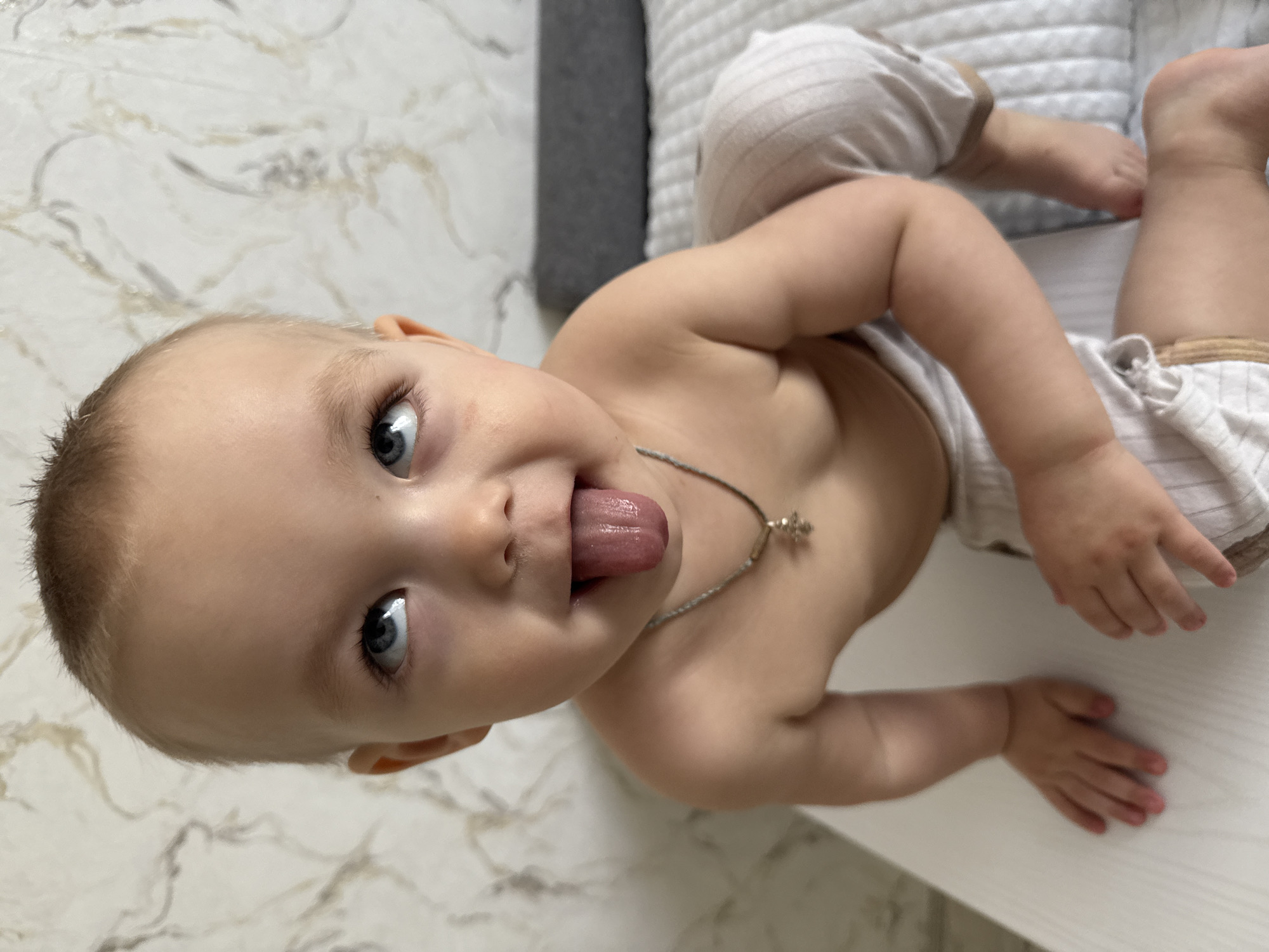 A baby lies on its back with its tongue sticking out. The baby is wearing a silver necklace with a cross pendant and striped white pants. The baby is resting on a white surface, possibly a table or changing pad. The background includes a marbled surface and a gray and white patterned cushion. The location is Unknown, Unknown.