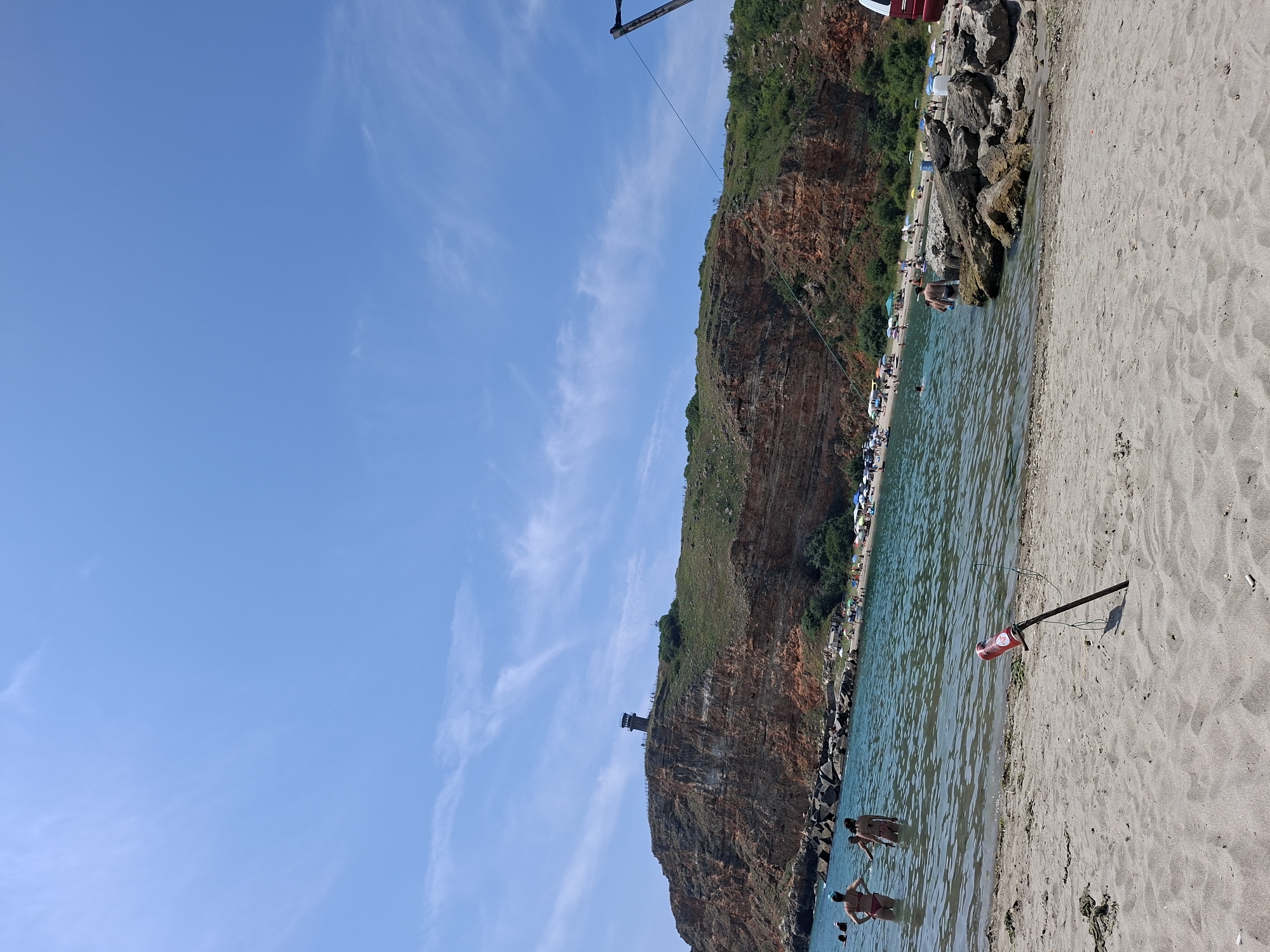 The image depicts a scenic coastal view of the city of Balgarevo, Bulgaria, on a clear, sunny day. The foreground is a sandy beach with footprints and some scattered seaweed. In the mid-ground, the clear blue sea is populated with several individuals swimming and wading.
Dominating the background is a dramatic, red-hued cliff face, characteristic of the Bulgarian Black Sea coast, topped with lush green vegetation. A prominent tower-like structure, likely a lighthouse or observation point, stands atop the cliff, offering panoramic views of the surroundings.
Along the shoreline at the base of the cliff, a beach with numerous umbrellas and sunbathers is visible. The water is calm, with gentle waves lapping the shore. A small, red object attached to a pole is planted in the sand near the water's edge in the foreground. The sky is a vibrant blue, with sparse, wispy white clouds. The overall atmosphere suggests a popular tourist destination, likely during the summer season.