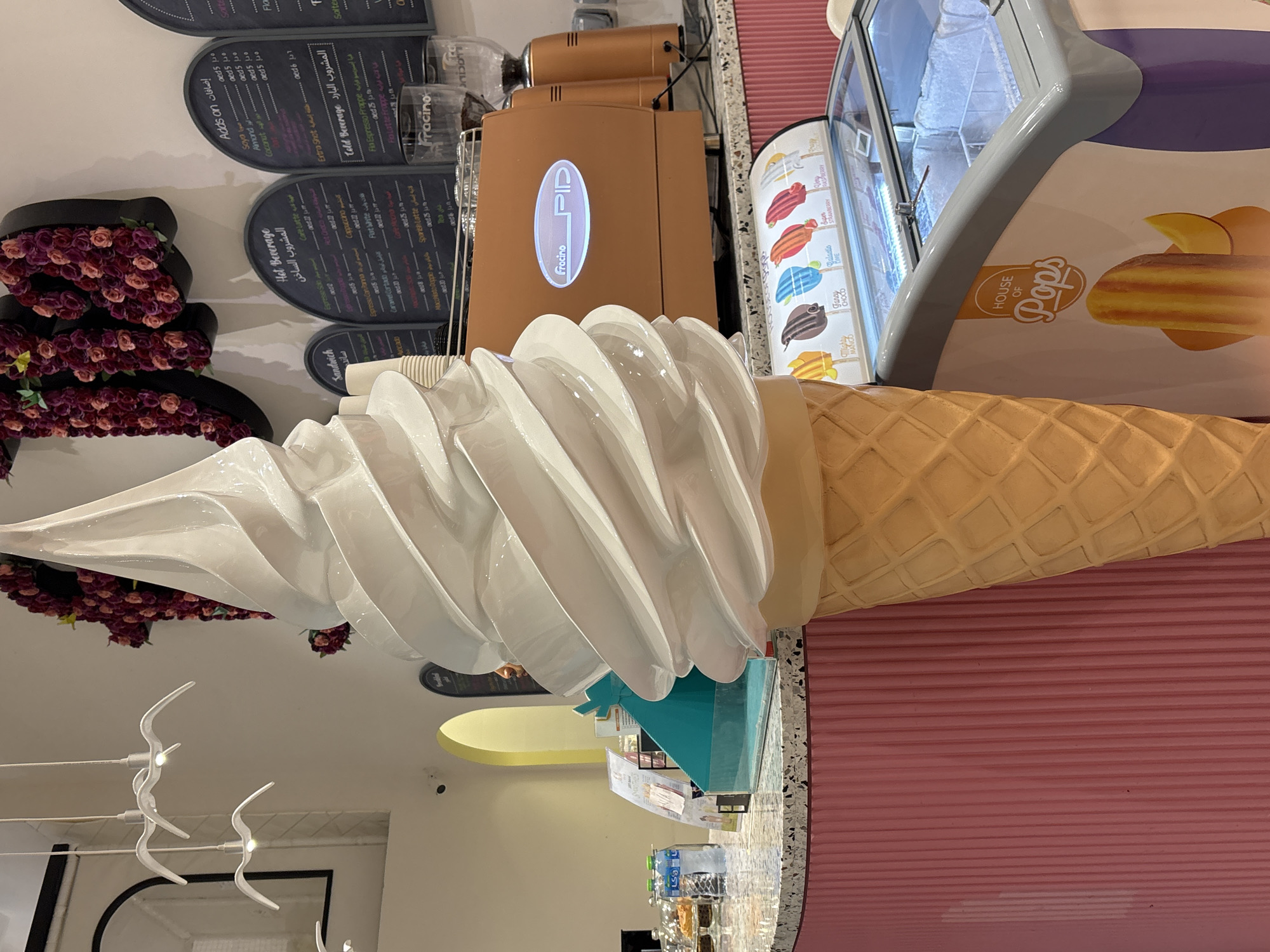 This is a full-shot, eye-level, indoor photograph of an ice cream shop in Dubai, United Arab Emirates. In the foreground, a large, sculptural, white soft-serve ice cream cone dominates the scene, tilted slightly to the right. Behind it, a pink, fluted counter runs across the bottom of the frame. On the counter, to the left of the ice cream sculpture, are several water bottles, a display case with pastries, and a teal-colored cash register. Behind the counter, to the right of the ice cream sculpture, is a metallic orange espresso machine with the brand "Fracino" visible. Further back, a menu board is mounted on the wall, displaying various hot and cold beverages. Above the menu board, a wall decoration of floral letters spelling "MO" is visible, composed of red and pink roses. Three white, bird-shaped pendant lights hang from the ceiling on the left side of the image. In the lower right corner, a glass-fronted freezer display case shows various popsicles with their names displayed, including "Mighty Mango," "Fancy Choco," "Galactic Lime," "Super Strawberry," and "Very Raspberry." The overall atmosphere is bright and inviting, with a focus on ice cream and coffee offerings.