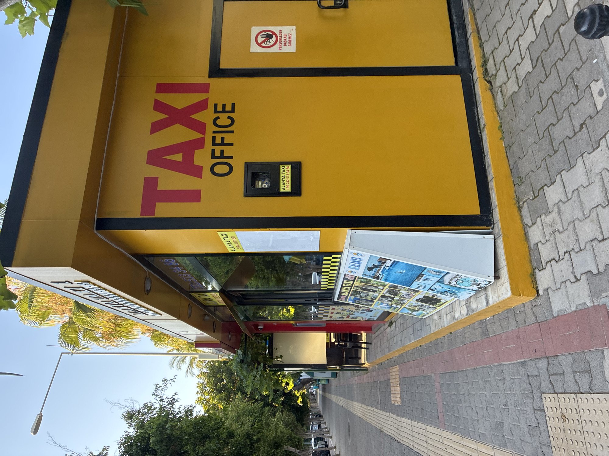 A bright yellow taxi office with "TAXI OFFICE" emblazoned in red letters on its facade is situated on a street in Alanya, Turkey. The office has a glass window displaying various promotional posters, likely for local attractions. To the left of the office, a sidewalk lined with trees stretches into the distance, with parked cars visible along the street. A street lamp stands tall on the sidewalk. On the right side of the taxi office, a closed door is marked with a "No Unauthorized Personnel" sign in Turkish. The ground is paved with interlocking bricks, with a red brick accent line running along the edge of the sidewalk. The scene appears to be captured during the daytime, with clear skies and natural light illuminating the area.