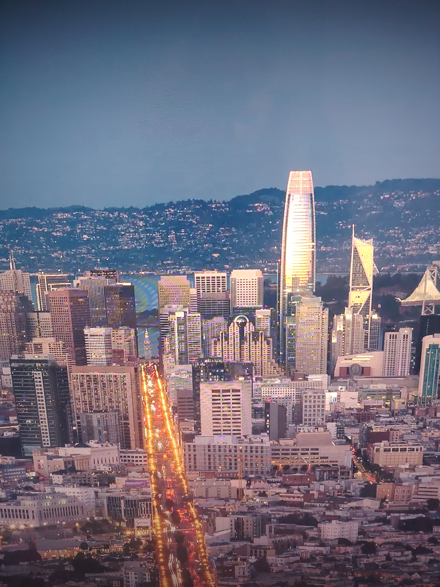 The image presents a high-angle view of a dense cityscape, likely San Francisco based on the architecture. A prominent, tall skyscraper with a reflective, elongated shape dominates the skyline, catching the light. Other buildings of varying heights and architectural styles surround it. A highway or major road cuts through the urban grid, illuminated by streams of car headlights and taillights, creating linear patterns of orange and red. In the background, a hilly landscape is visible under a hazy sky, adding depth to the scene. The overall lighting suggests either dusk or dawn.