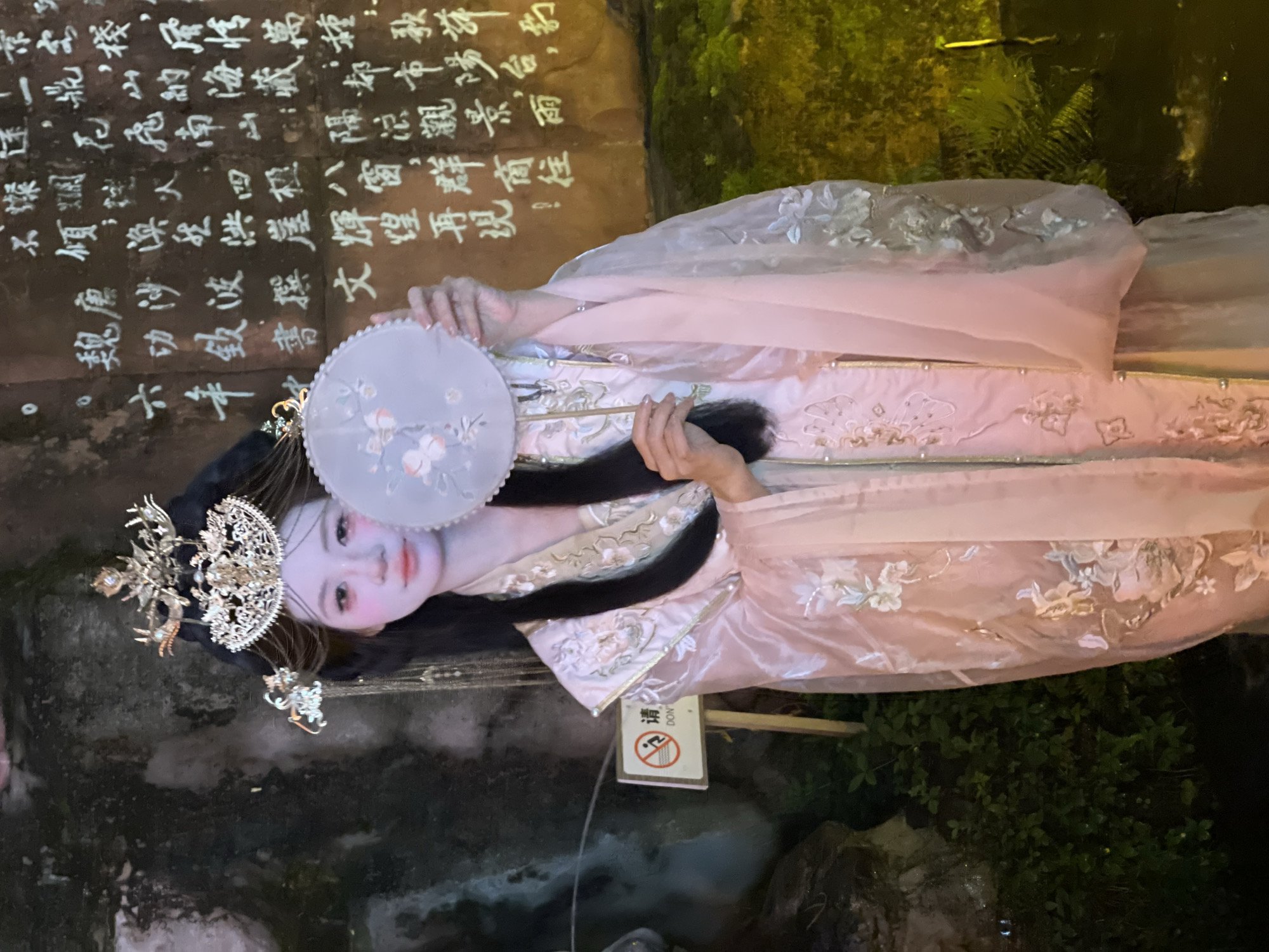 A female subject stands in an outdoor environment in Yuzhong District, China. She wears a light pink Hanfu-style ensemble featuring white floral embroidery and pearl accents. Her dark hair is styled in an elaborate updo, adorned with a large, ornate gold and silver headdress. Her face exhibits pale makeup with pronounced pink blush and red lips. She holds a light blue circular fan, decorated with white floral patterns, partially obscuring her lower face. To her left, a dark, textured rock wall is visible, with green foliage at its base and a white sign bearing a red "no diving" or "no climbing" symbol on a wooden post. To her right and rear, a light-colored rock wall is illuminated, displaying numerous white Chinese characters. Below this inscribed wall, a dark body of water is present.
