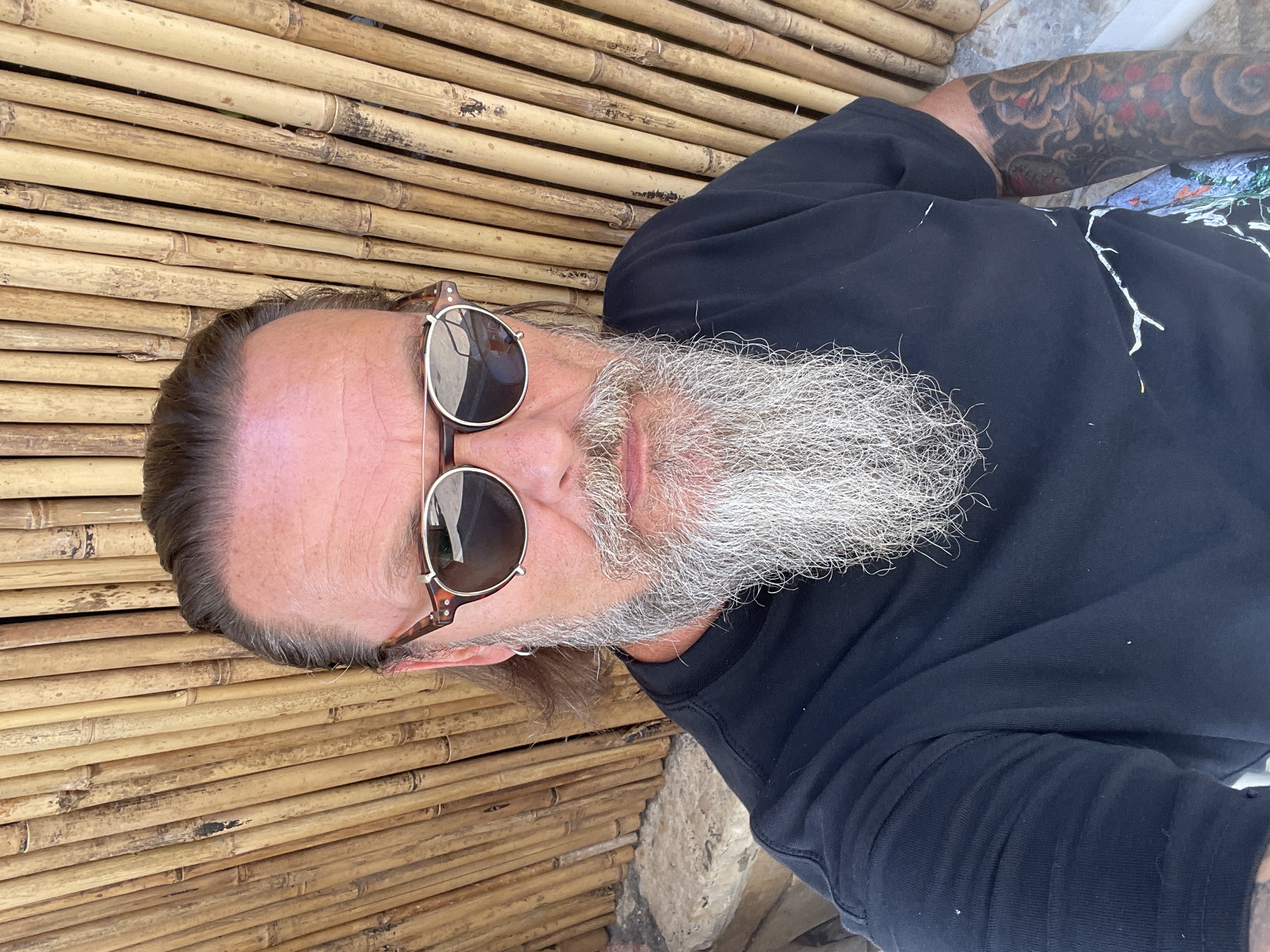 A man with a long, gray beard, wearing sunglasses and a black t-shirt, poses for a selfie in front of a bamboo wall. He has tattoos on his left arm. The image was taken on a sunny day. The location is in the city of Netanya, Israel.