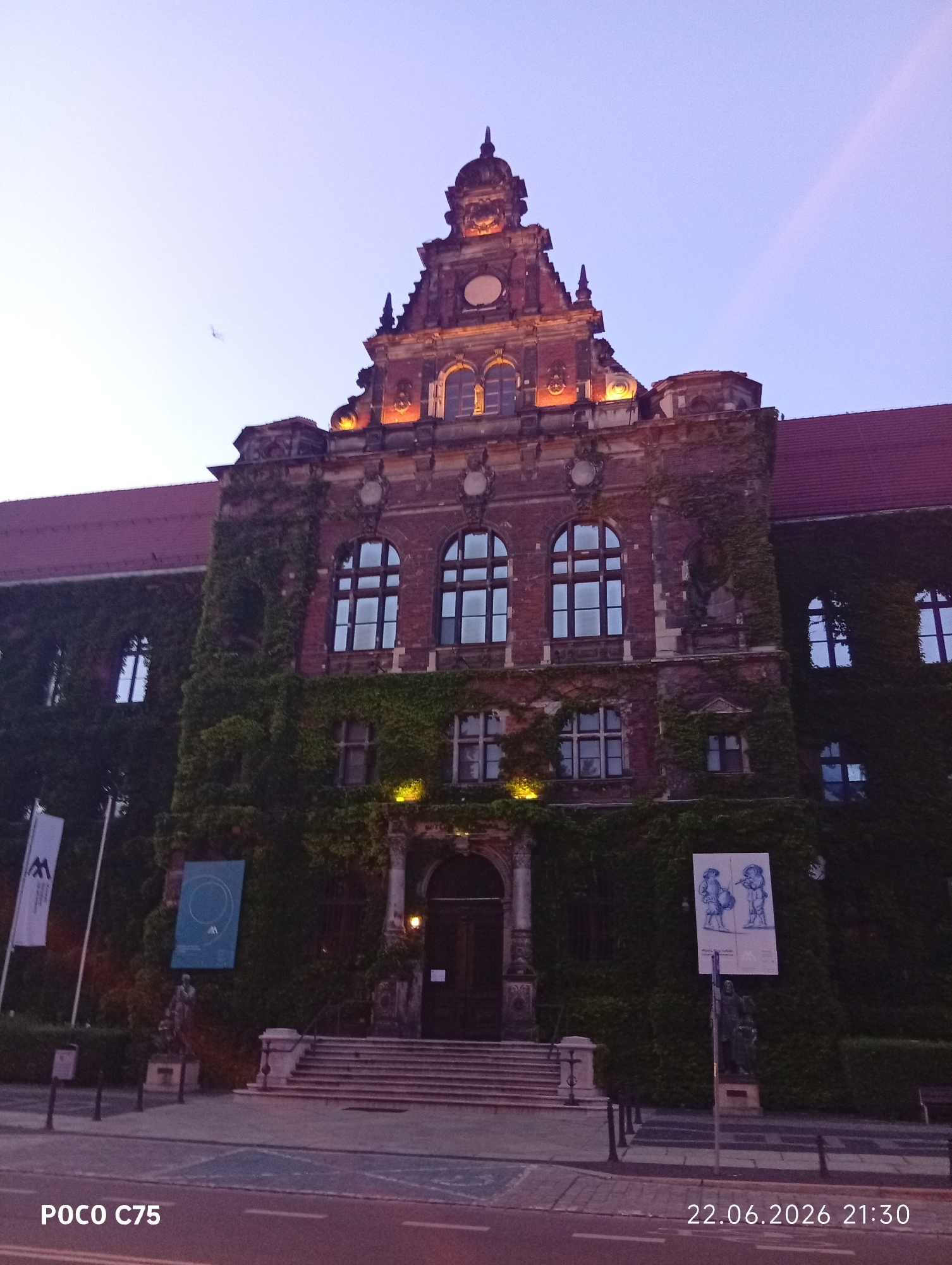 The image shows the front of a large, old, brick building covered in ivy. The building has a prominent, ornate facade with multiple arched windows and decorative architectural elements, including a tower-like structure at the top. The windows are lit from within, casting a warm glow. There are stairs leading up to the main entrance, which is flanked by statues and large posters. Flags are also visible on the left side of the building. The sky is a pale, twilight blue. The photograph includes a timestamp in the bottom right corner reading "22.06.2026 21:30" and a watermark "POCO C75" in the bottom left corner. This building is located in Wrocław, Poland and is identifiable as the Faculty of Chemistry building at the University of Wrocław, situated at Joliot-Curie street.